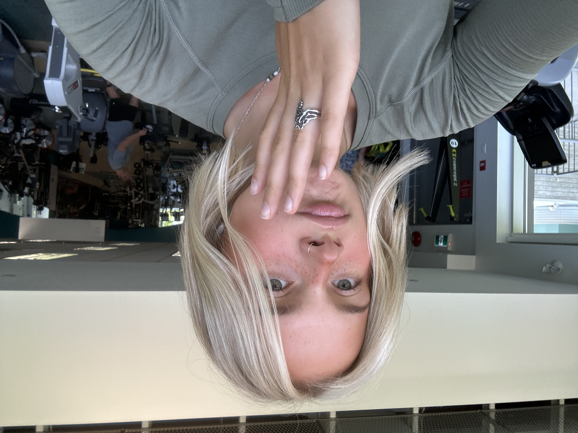 A young woman with blonde hair and freckles, wearing a green long-sleeved shirt and a silver ring, is taking a selfie in a gym. She is holding her left hand up to her chin, with her fingers spread. In the background, there are exercise machines, including treadmills and rowers. A man in a grey t-shirt and black shorts is visible on a treadmill in the distance. The gym appears to be indoors, with large windows looking out onto an outdoor area with trees. The overall lighting is bright, suggesting it is daytime. There is no indication of the location in Whistler, Canada.