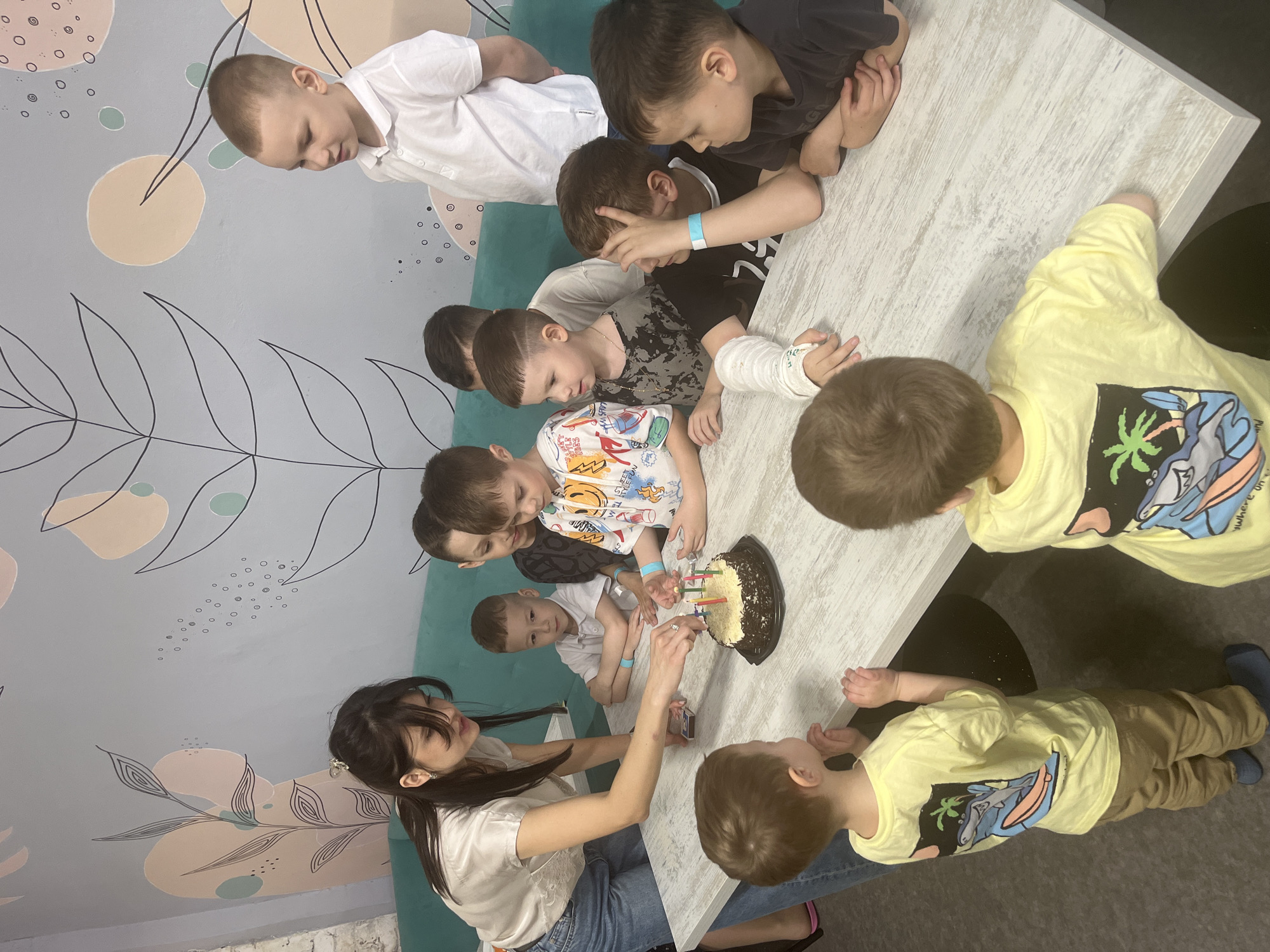 A group of approximately ten children are gathered around a rectangular table. A woman in a white shirt and jeans is lighting candles on a chocolate cake. One child has a cast on their arm. The location appears to be a decorated interior with floral and geometric patterns on the wall. This is likely a birthday celebration. The location is Ulyanovsk, Russia.