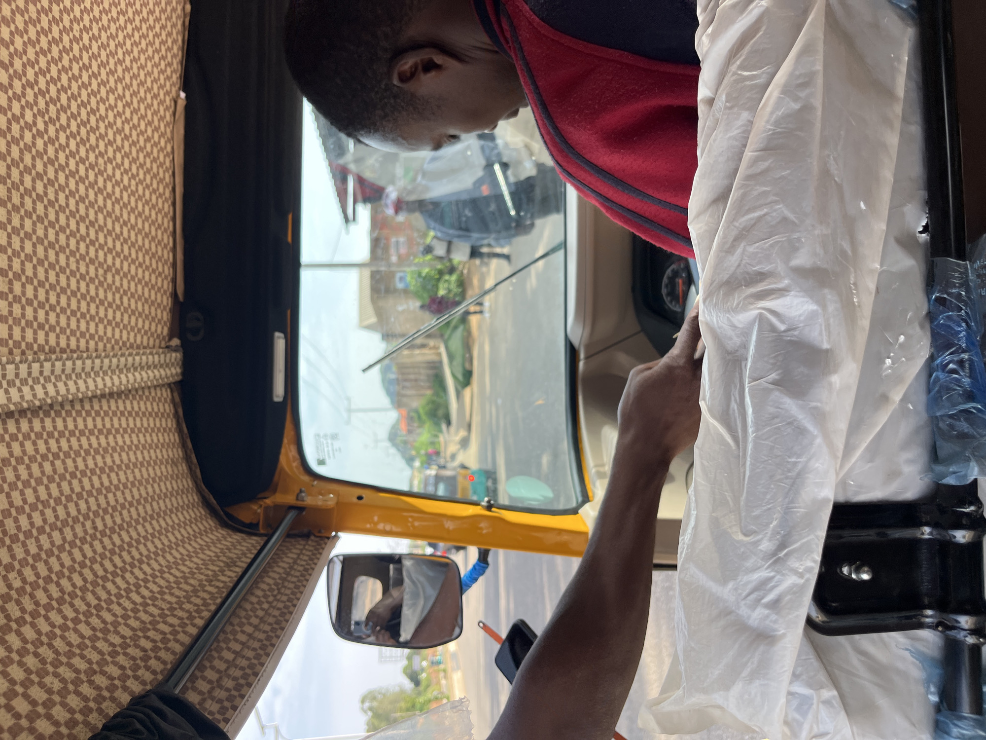 The image captures the interior of a yellow auto-rickshaw, likely in Bukuru, Nigeria, viewed from the passenger's perspective. A person wearing a red and black garment is seated in the driver's position, facing forward, their head turned to the right. Their arm and hand are visible on the right side of the frame, resting near the steering mechanism, which is covered by white plastic sheeting. Another arm and hand, belonging to someone in the passenger area, are extended towards the front of the vehicle, touching the white plastic covering. The auto-rickshaw's interior features a checkered brown and beige fabric lining on the ceiling. Through the windshield, a street scene unfolds, showing other vehicles, buildings, and pedestrians in the distance. The road appears to be paved, and the surroundings suggest an urban or semi-urban environment. The overall lighting indicates it is daytime, with sunlight illuminating the scene.