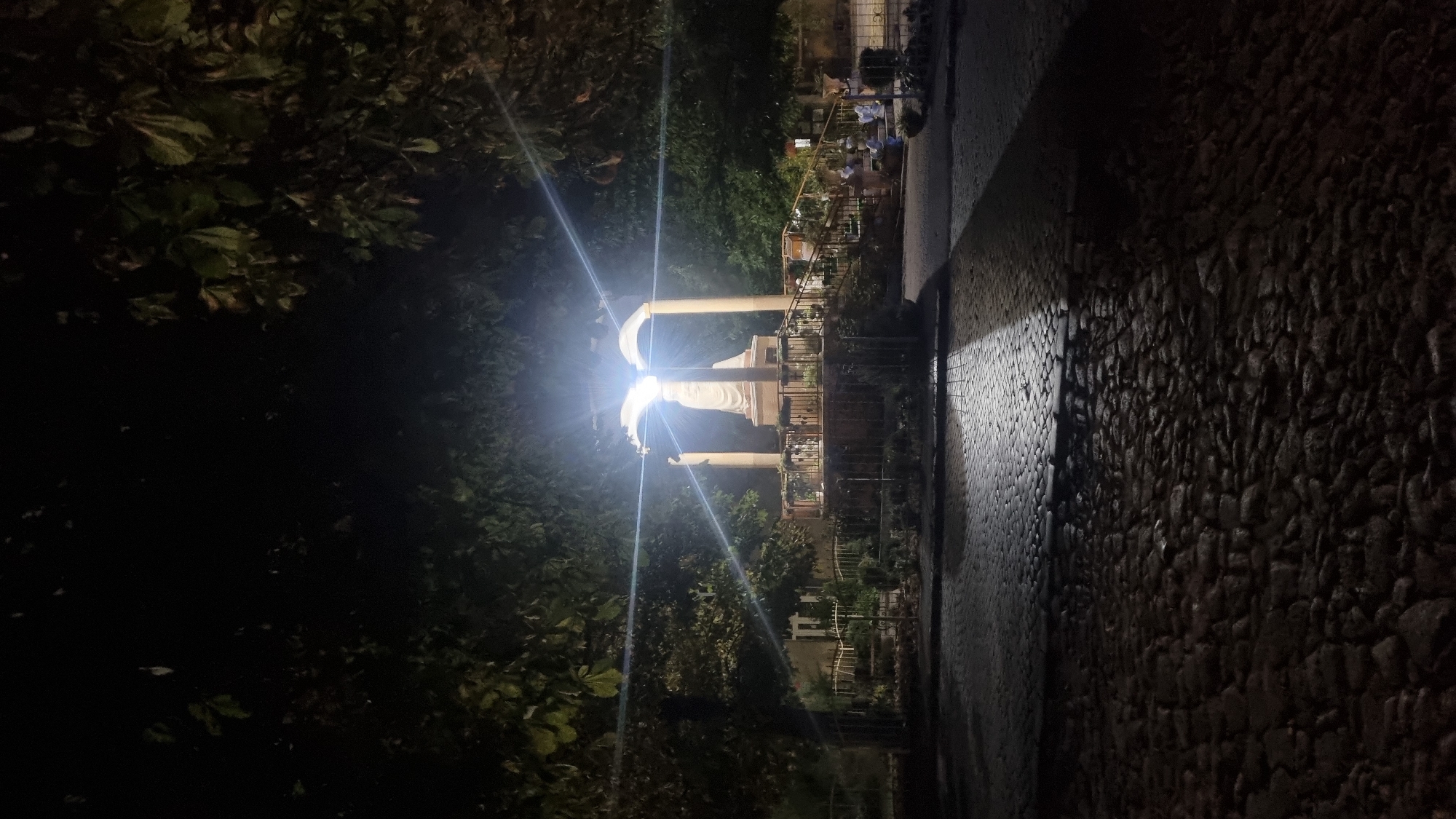 A statue of Mary is illuminated by bright lights under an archway at night. The statue is located in a public square in Berehove, Ukraine. The square is paved with cobblestones and surrounded by trees. Several people are visible in the background, some sitting on benches and others walking. The scene is peaceful and serene.