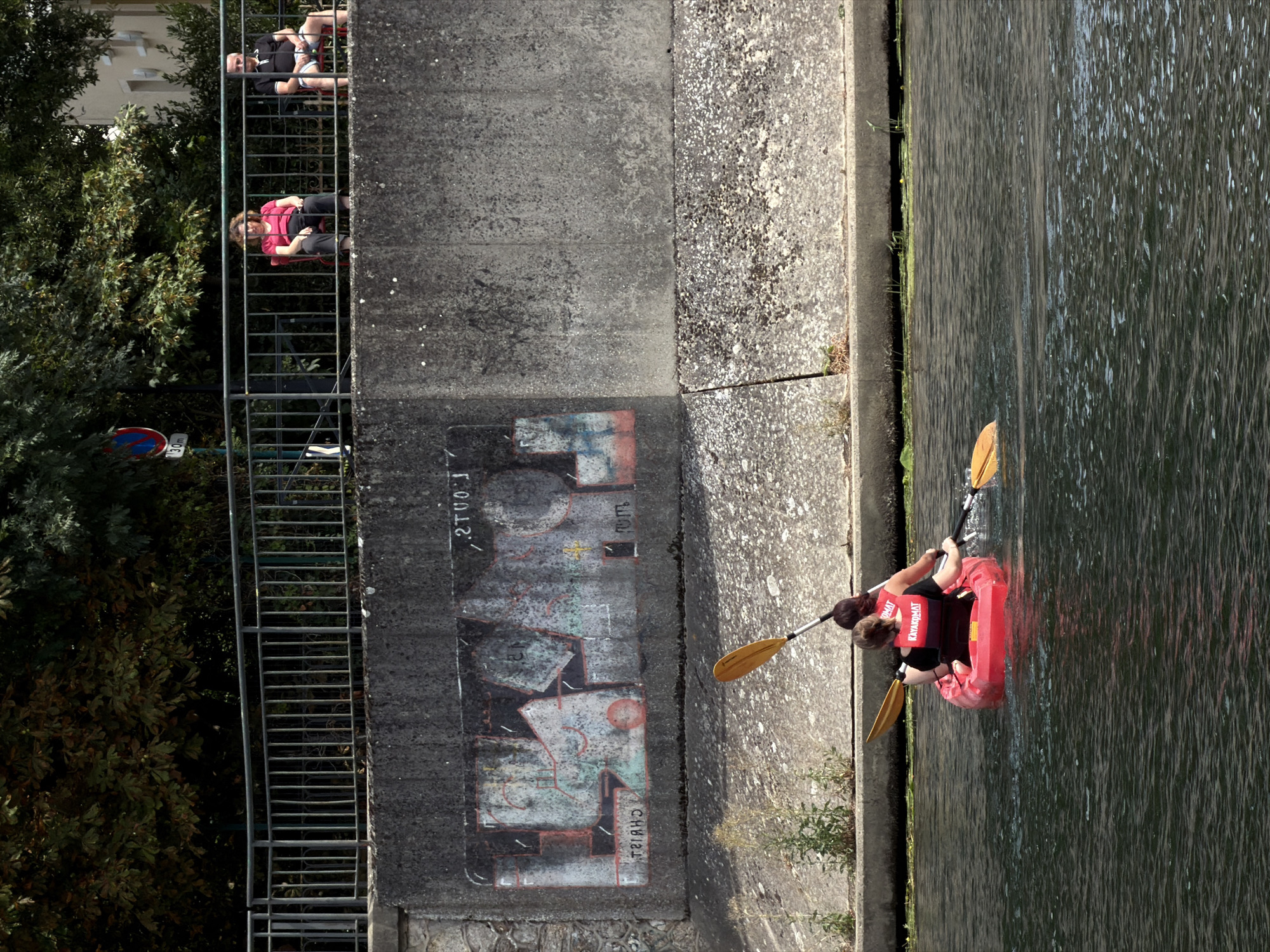 The image depicts a canal scene in Joinville-le-Pont, France, likely the Marne River due to its characteristic canalization and bordering parkland. Two individuals are kayaking in a red kayak on the dark, rippling water. The person in front, wearing a life vest with "KAYAKMAT" printed on it, is facing away from the camera and paddling. The person behind them is also facing away and appears to be paddling as well.
Above the canal, a concrete embankment features graffiti artwork. Beyond this, a metal railing runs along the top of the embankment. On the other side of the railing, on a paved area, two people are seated on chairs. A woman in a pink top is seated on a red chair, and a man in a black polo shirt and blue shorts is on a white chair to her right. Trees with green and brown foliage are visible behind them. A traffic sign with a red circle and a diagonal line, and "30m" written on it, is also visible to the left of the seated individuals. The overall scene suggests a sunny day with the individuals enjoying recreational activities.