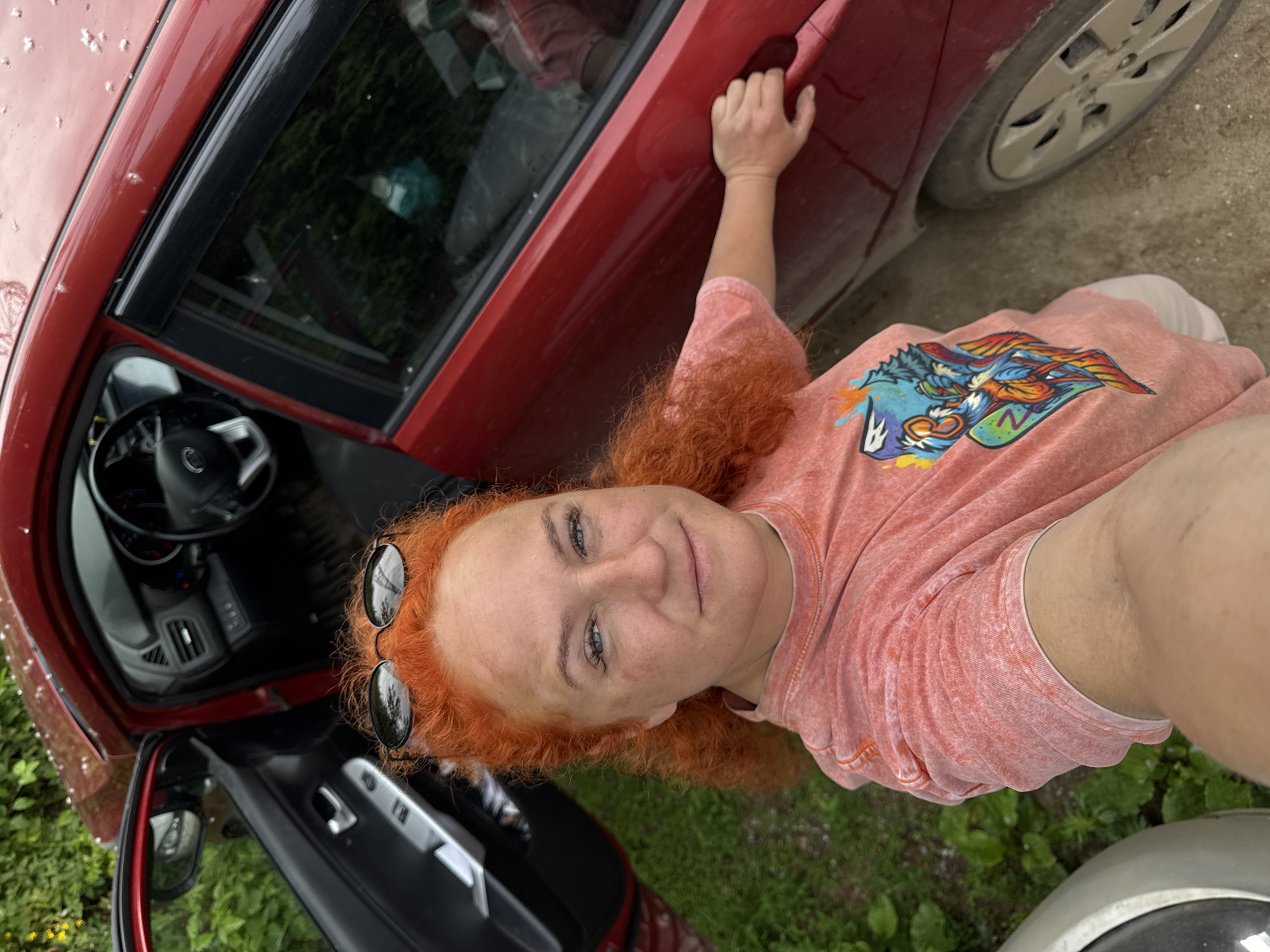 A Caucasian woman with bright orange hair, wearing sunglasses on her head and a coral-colored t-shirt with a colorful graphic, is standing next to a red car with the driver's side door open. She is holding the car door handle with her right hand and appears to be taking a selfie. The interior of the car, including the steering wheel and dashboard, is visible. The environment appears to be outdoors, with green foliage and a gravelly surface. The location is identified as Chaikovsky, Russia.