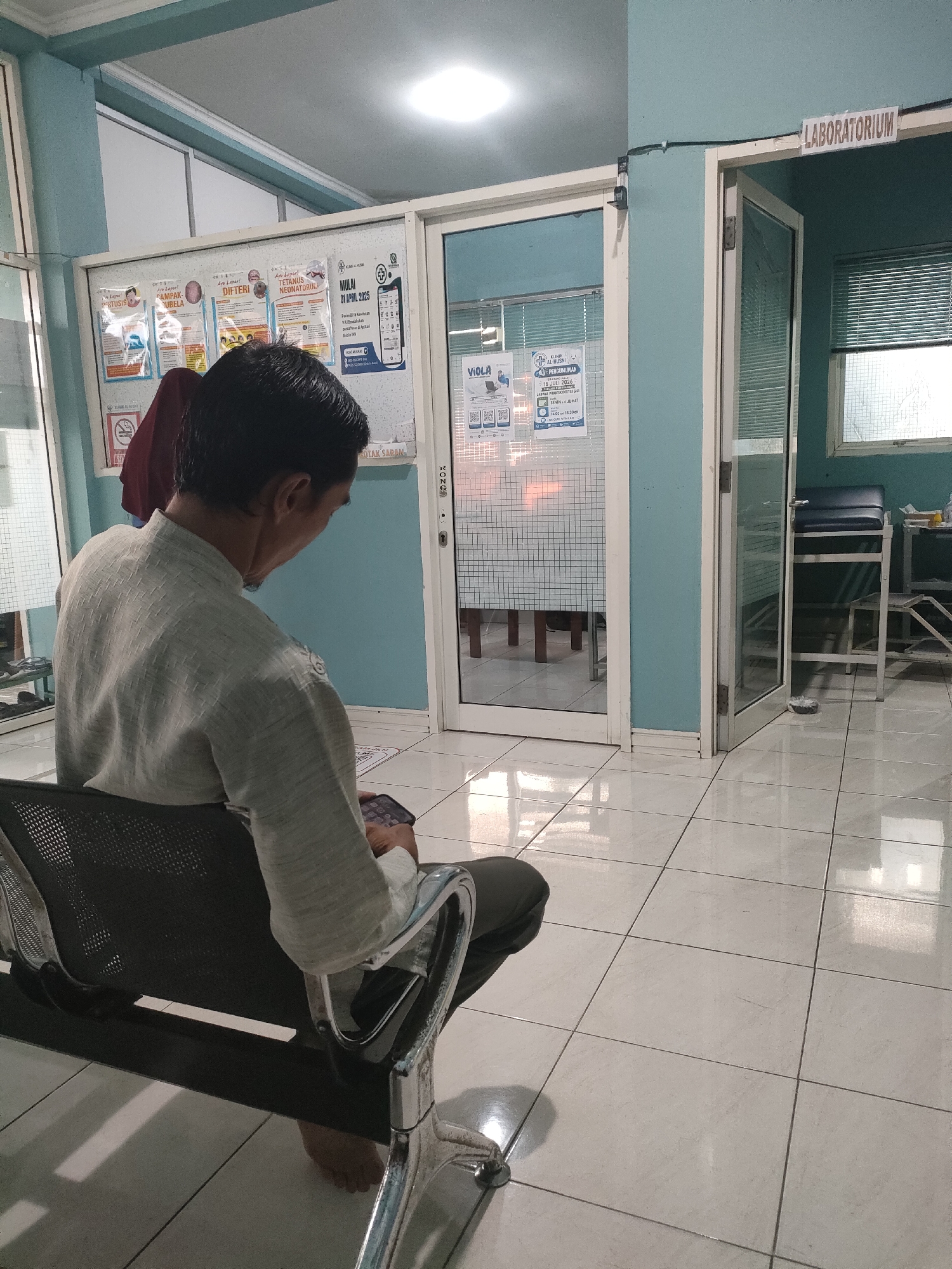 A medium shot shows a man sitting in a waiting area. The man is seated on a black chair with a metal frame, facing away from the camera. He is wearing a light-colored, long-sleeved shirt and dark pants, and holding a mobile phone in his hands. The man appears to be looking at the phone. The waiting area is indoors, with pale blue walls and a tiled floor. To the man's left, there is a glass-paneled door with a notice board on the wall next to it, displaying several informational posters. To the man's right, there is another glass-paneled door with a sign above it reading "LABORATORIUM". Through this door, a chair and a metal table are visible. The overall lighting is bright, with overhead fluorescent lights visible on the ceiling. The scene suggests a clinic or hospital waiting room. The sign "KLINIK AL-HUSNI" is visible on one of the posters. This clinic is located in Sungailiat, Indonesia.