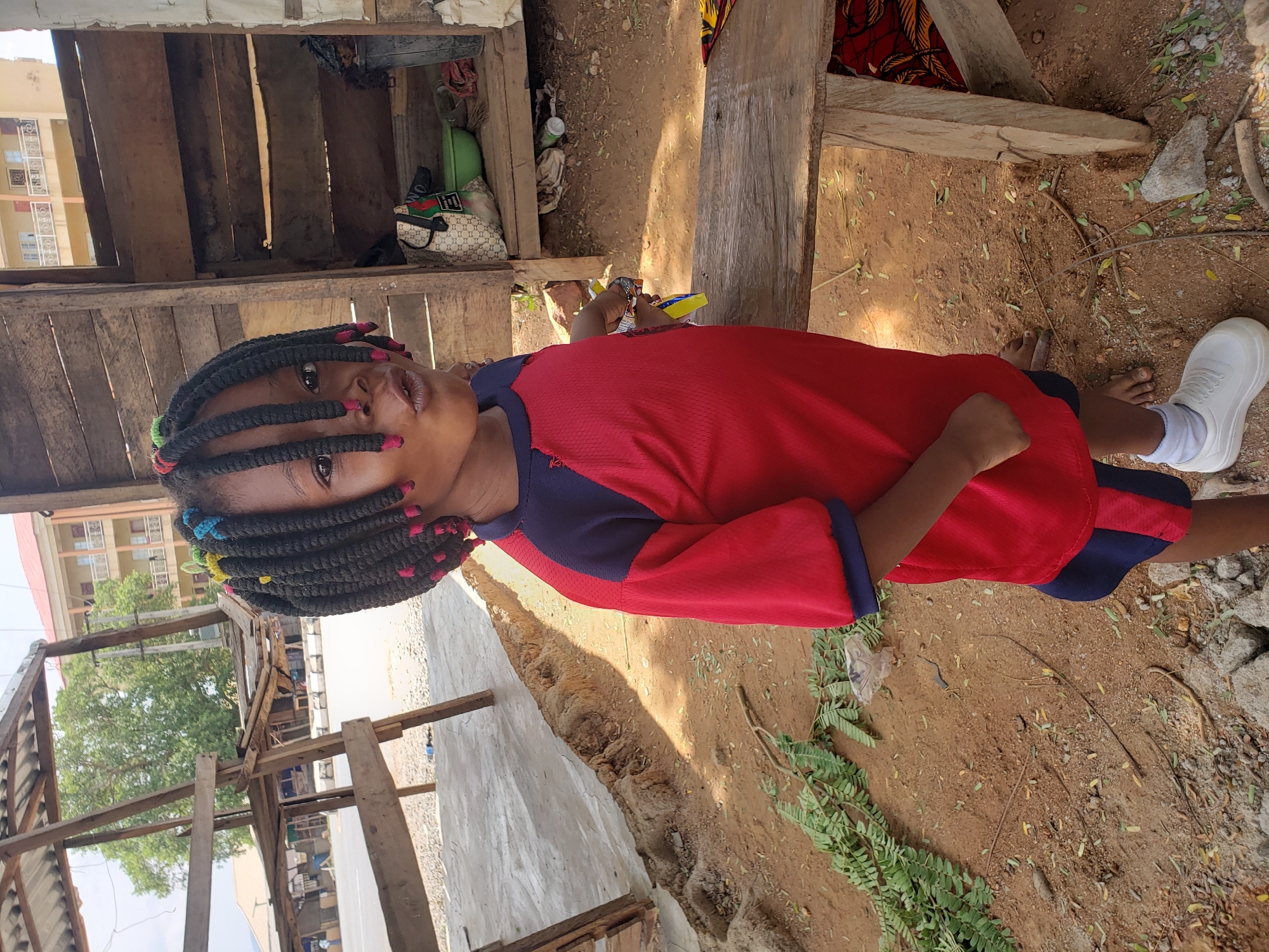 A young person stands in a dirt area in Adankolo, Nigeria. The individual wears a red and blue shirt and white socks and a white shoe on one foot. The person has dark braids with colorful embellishments. The ground is brown dirt mixed with green vegetation. The person is positioned near wooden structures, including a shack-like building with items stored inside, such as bags and containers. In the background, a multi-story building is visible.