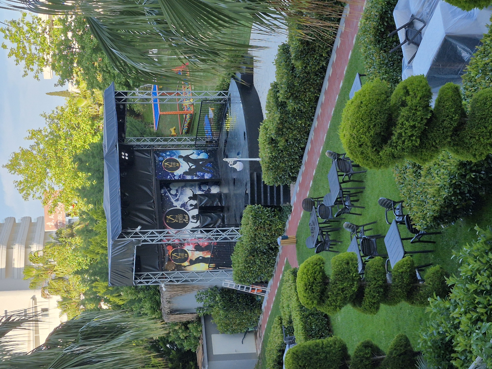 A ground-level, outdoor view of a stage in Alanya, Turkey. The stage is covered in black tarps and displays the logo of "Diamond Hotels." The surrounding landscaping includes a brick-laid path, manicured shrubbery, and tables with chairs placed on the grass. A playground area is visible in the background. Palm trees and other greenery surround the area, suggesting a tropical or subtropical setting. An unidentified building with a sign "FABRIC" can be seen partially in the background.