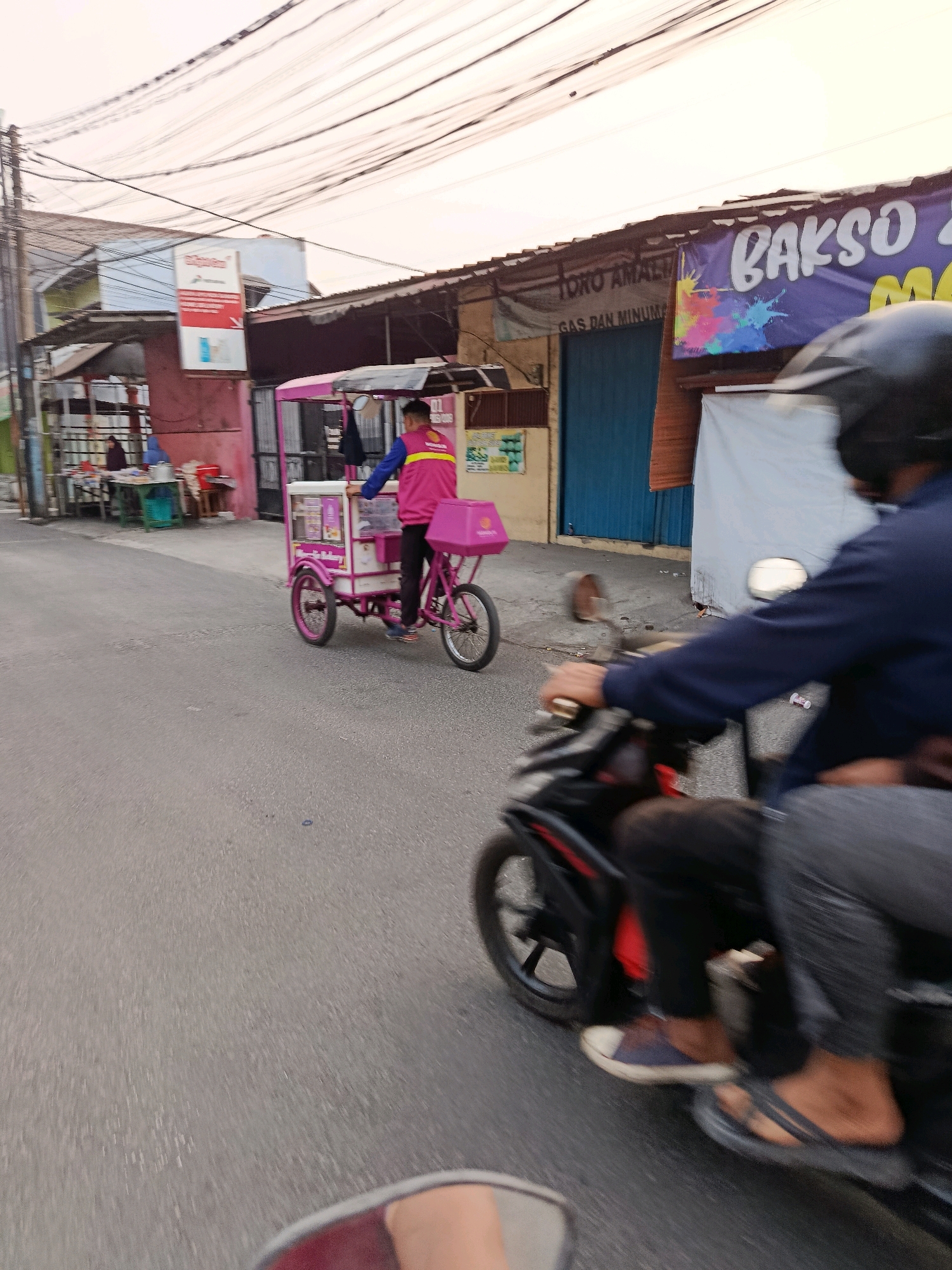 The image captures a street scene in Bekasi, Indonesia. A person on a motorcycle is in the foreground, seen from behind, wearing a blue jacket and black pants. The motorcycle is black with red accents. In the middle ground, an ice cream cart, pink and white, is being pushed by a person wearing a pink vest over a blue shirt and black pants. The cart has several compartments and a pink box on the back. Behind the ice cream cart, there are small shops with awnings and signs, one with the word "BAKSO" visible. The street is paved, and power lines are strung overhead. The sky is overcast with a pale orange hue. The overall impression is a busy, everyday street scene in an urban area.