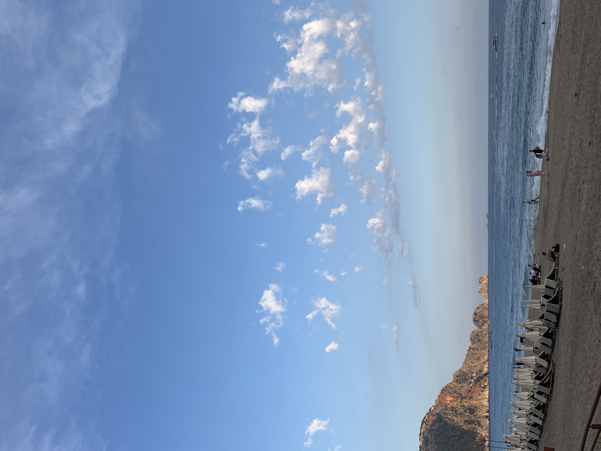 The image shows a beach in Alanya, Turkey. The view is oriented vertically, with the blue sky and scattered cumulus clouds taking up the majority of the frame. The Mediterranean Sea is visible along the right side of the image, with a gentle surf breaking on the sandy beach. People are present on the beach, both near the water and further up on the sand. Rows of beach chairs and umbrellas are set up, suggesting a resort or tourist area. A rocky headland or small mountain rises out of the sea near the beach. The lighting suggests it is daytime.