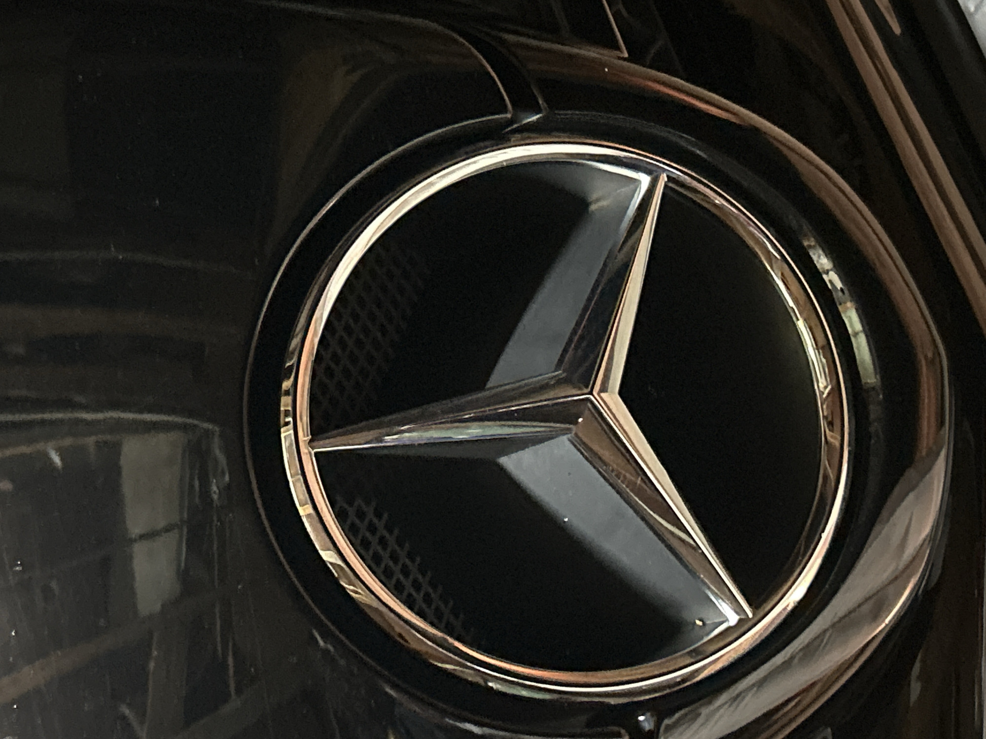 The image shows a close-up of a Mercedes-Benz emblem, specifically the three-pointed star enclosed in a circle, mounted on what appears to be a black car grill. The star and circle are silver/chrome in color, contrasting with the black background of the grill. The emblem is slightly off-center. No people, actions, or interactions are visible in the image. The location is not identifiable from the content of the image. The image is a close-up of a Mercedes-Benz emblem. There is nothing to suggest that this image was taken in the city of Sofia, Bulgaria.