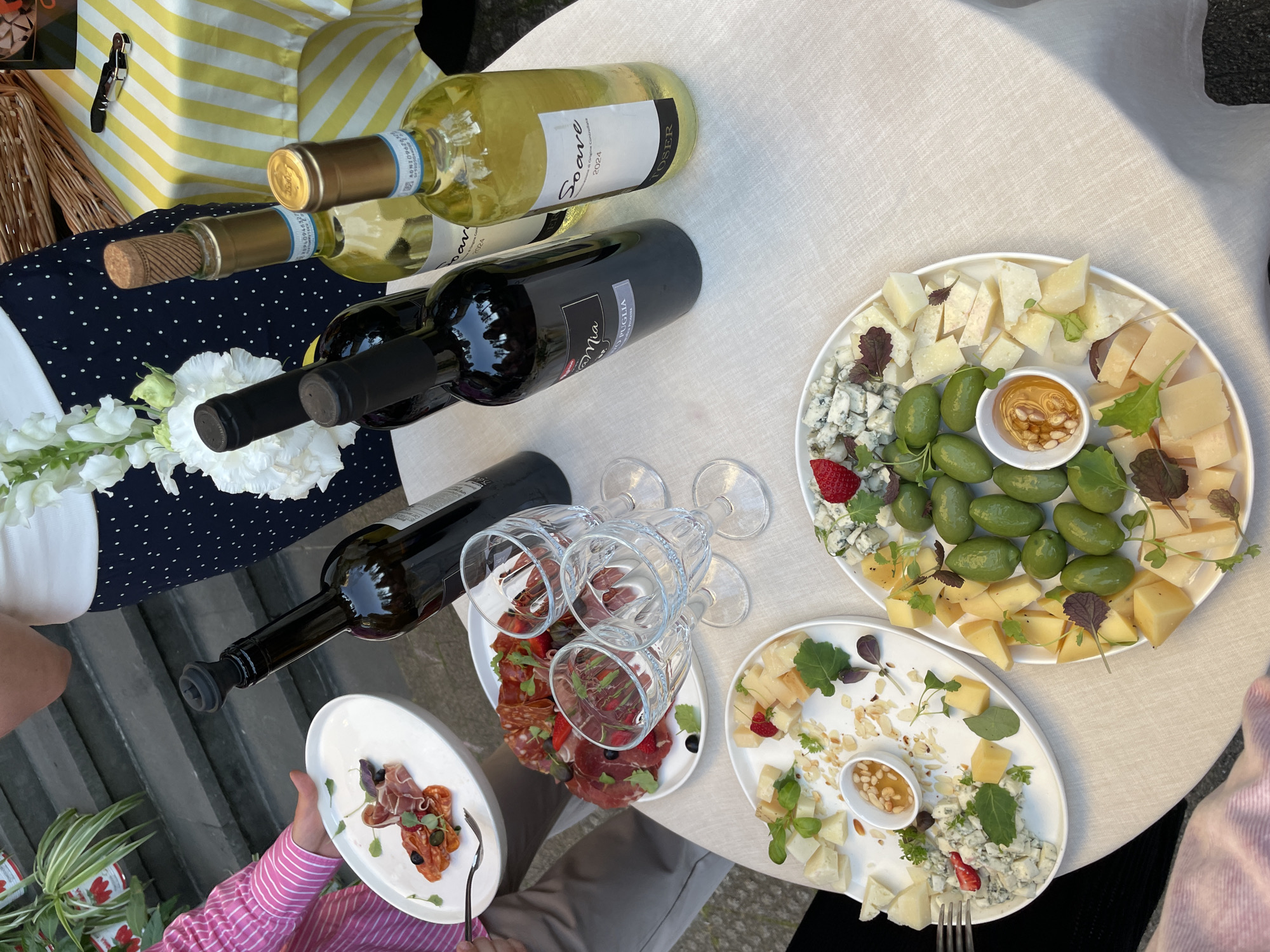 A close-up overhead shot captures a table laden with food and drinks, set against a backdrop suggesting an outdoor gathering. The table is covered with a light-colored, textured tablecloth. Prominently featured are three plates of appetizers. The first plate, positioned on the left, holds sliced cured meats, capers, and microgreens. The second, more central plate, displays a medley of sliced cured meats, garnished with strawberries and microgreens. The third plate, to the right, presents a selection of cheeses, olives, and a small dish of honey with nuts, also adorned with strawberries and microgreens.
Several bottles of wine are visible. Two dark-colored bottles stand upright, one with a black stopper, and another with a cork. Two lighter-colored wine bottles are also present, both with corks. Three empty wine glasses are placed between the appetizer plates and the wine bottles, suggesting recent use. In the background, a person wearing a pink striped shirt is partially visible, holding a plate. Another individual is seen wearing a dark blue skirt with white polka dots and a white top. The environment appears to be outdoors, possibly a garden or patio, with steps and foliage visible in the upper left corner. A yellow and white striped cloth is visible in the background on the right. The overall impression is of a relaxed social event with a focus on food and wine.