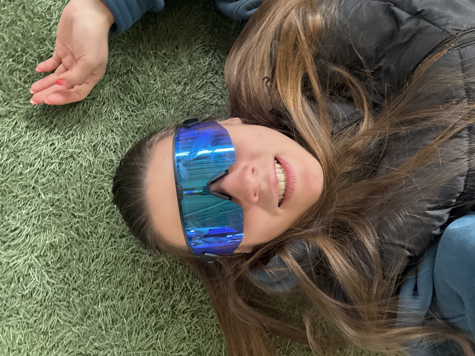 A person is lying on a green, grassy surface. The person is wearing large, blue mirrored sunglasses. Their hair is brown and spread around their head. The person is smiling. A hand is visible in the upper left corner of the image, with red nail polish on some of the nails. The person is wearing a black puffer vest and a blue top. The location is Unknown, Unknown.