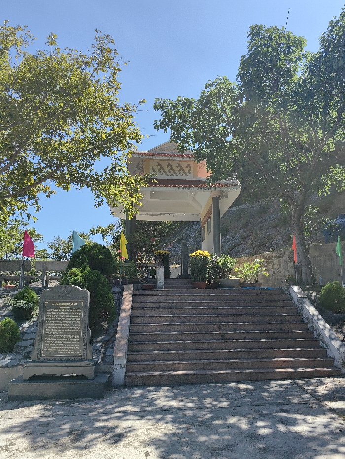 A sunny day illuminates a building with a distinct roof structure, surrounded by lush greenery and trees. A set of wide stairs leads up to the building, flanked by a stone monument on the left. Colorful flags are positioned on the sides, adding vibrancy. The scene evokes a peaceful and possibly historical location.