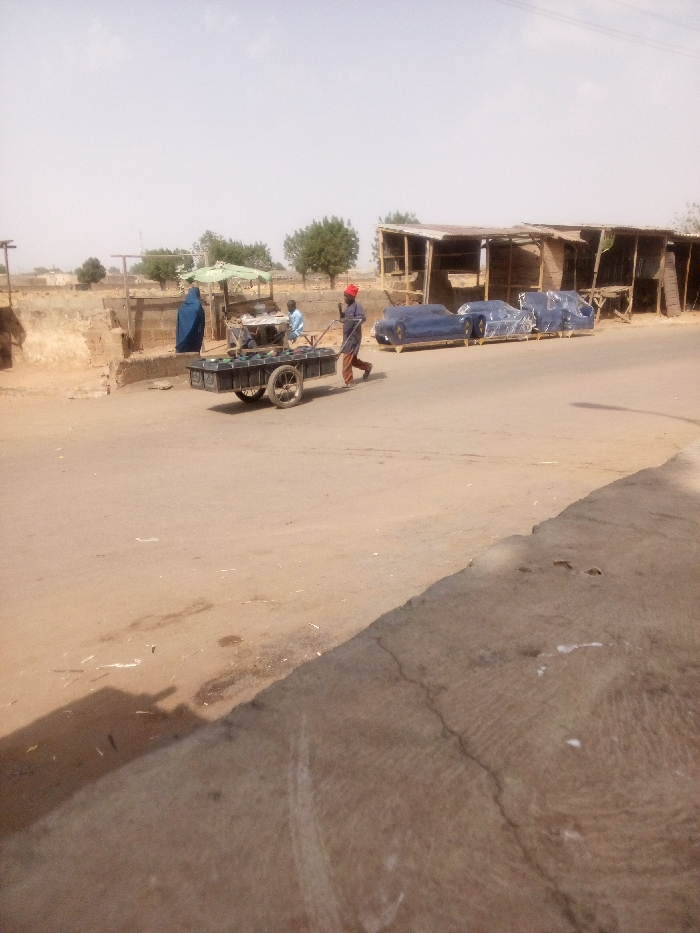 This image captures a daytime street scene in Tudun Murtala, Nigeria. The scene is dominated by a wide, unpaved or dirt road that extends into the background.
In the mid-ground, a male individual, dressed in a red cap, dark long-sleeved shirt, and light-colored trousers, is actively pushing a dark-colored, two-wheeled handcart towards the right side of the frame. The cart is loaded with multiple green, cylindrical objects, likely bottles or containers, arranged in rows.
To the left of the road, behind the handcart, a market area is visible. A female figure, clad in a full blue garment, stands near a light-colored building or stall. Adjacent to her, a light green market umbrella shades a stall where another individual is seated, with a third person partially visible nearby. The background on the left consists of low, earth-toned structures or walls and scattered green trees under a pale, hazy sky.
On the right side of the road, a series of rudimentary market stalls are constructed from wood frames and topped with corrugated metal or tarp roofs. In front of these stalls, three pieces of furniture, appearing to be sofas or chairs, are positioned along the roadside. Each furniture item is distinct and fully wrapped in blue protective plastic, resting on what appear to be wooden pallets or bases.
The immediate foreground on the right features a cracked concrete curb or raised edge. Shadows are cast by people and objects, indicating sunlight from the upper left.