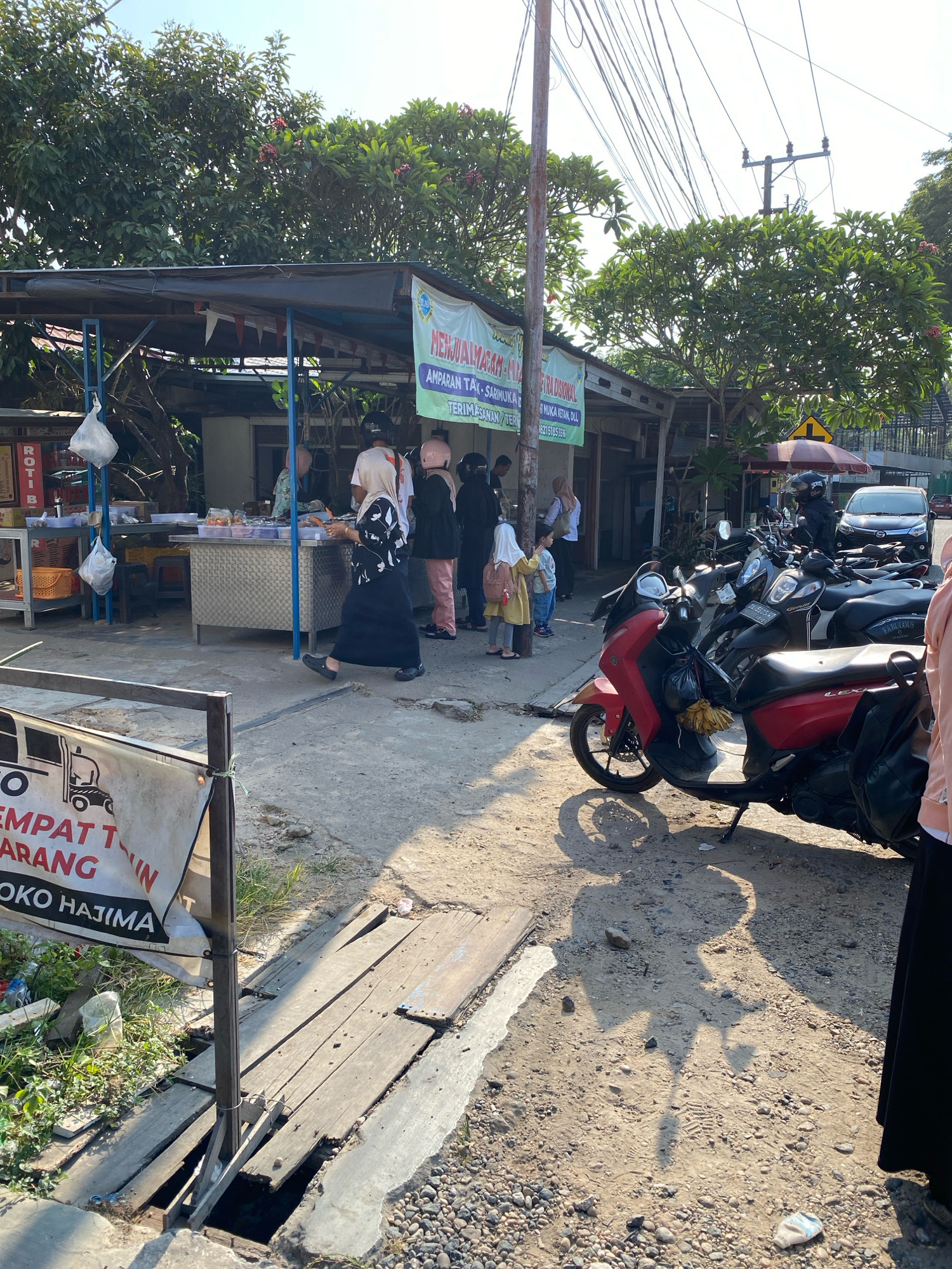 The image depicts a street scene in Banjarbaru, Indonesia, likely a market or roadside stall area. In the foreground, a red scooter is parked on a dirt and gravel surface, casting a distinct shadow. Behind it, several other motorcycles are lined up. A banner with Indonesian text, partially visible, hangs from a pole in the left foreground.
To the right, a row of roadside stalls is set up under a canopy. People are visible interacting with the stalls and each other. One person, wearing a black abaya and hijab, is walking away from the stalls. Another person, wearing a light-colored hijab and black and white patterned clothing, is standing and looking towards the stalls. Further back, other individuals in various attire, including headscarves, are present. The stalls appear to be selling food items, with displays of packaged goods and what look like snacks.
The environment is characterized by a sunny day with clear skies. Trees with green foliage and some pink flowers are visible behind the stalls. Electrical wires crisscross the upper portion of the image, typical of urban or semi-urban settings. The ground is a mix of compacted dirt, gravel, and some areas with wooden planks laid over what appears to be a drain or ditch. The overall impression is of a busy, everyday scene in a commercial area of Banjarbaru.