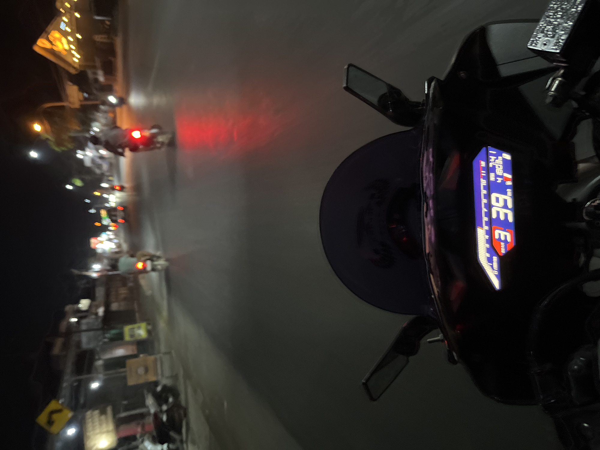 This is a first-person perspective shot from a motorcycle at night on a street in Kalinyamatan, Indonesia. The road is paved and appears wet from rain, reflecting the taillights of other vehicles and streetlights. In the foreground, the motorcycle's digital dashboard displays the gear (3), speed (39 km/h), and total mileage (41803). The dashboard also shows a bar graph indicating fuel level. The windshield of the motorcycle is visible, as are the side mirrors.
Further down the road, several motorcycles are visible, with riders illuminated by streetlights and their own vehicle lights. One motorcycle in the middle ground has its red taillight prominently glowing. The surrounding environment features buildings with illuminated signs and storefronts, typical of a commercial area at night. Streetlights are also visible, casting pools of light on the road and the sides of the street. A yellow traffic sign with a curved arrow is visible on the left side of the road. The overall scene depicts a typical urban nighttime commute or ride.