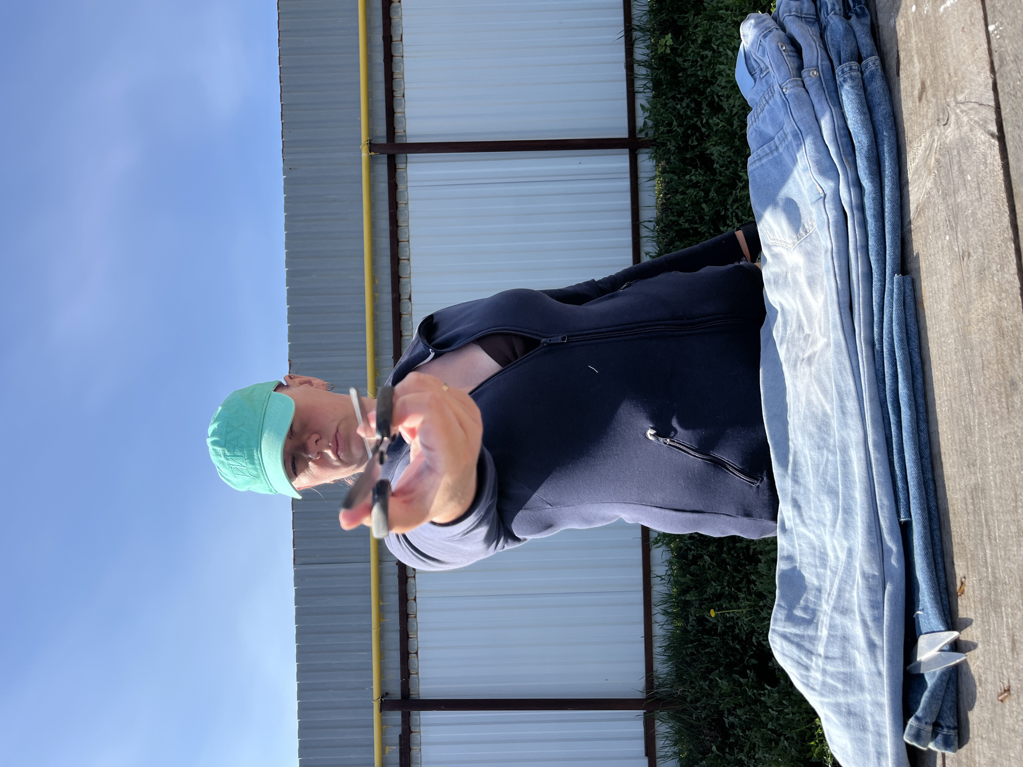A woman wearing a turquoise baseball cap and a dark blue zip-up sweatshirt is holding a pair of scissors in her right hand. She is standing near a stack of blue jeans on a weathered wooden surface. Behind her is a metal fence with a yellow support beam. In the background, there is green foliage and a clear blue sky. This image was taken in the city of Chaikovsky, Russia.