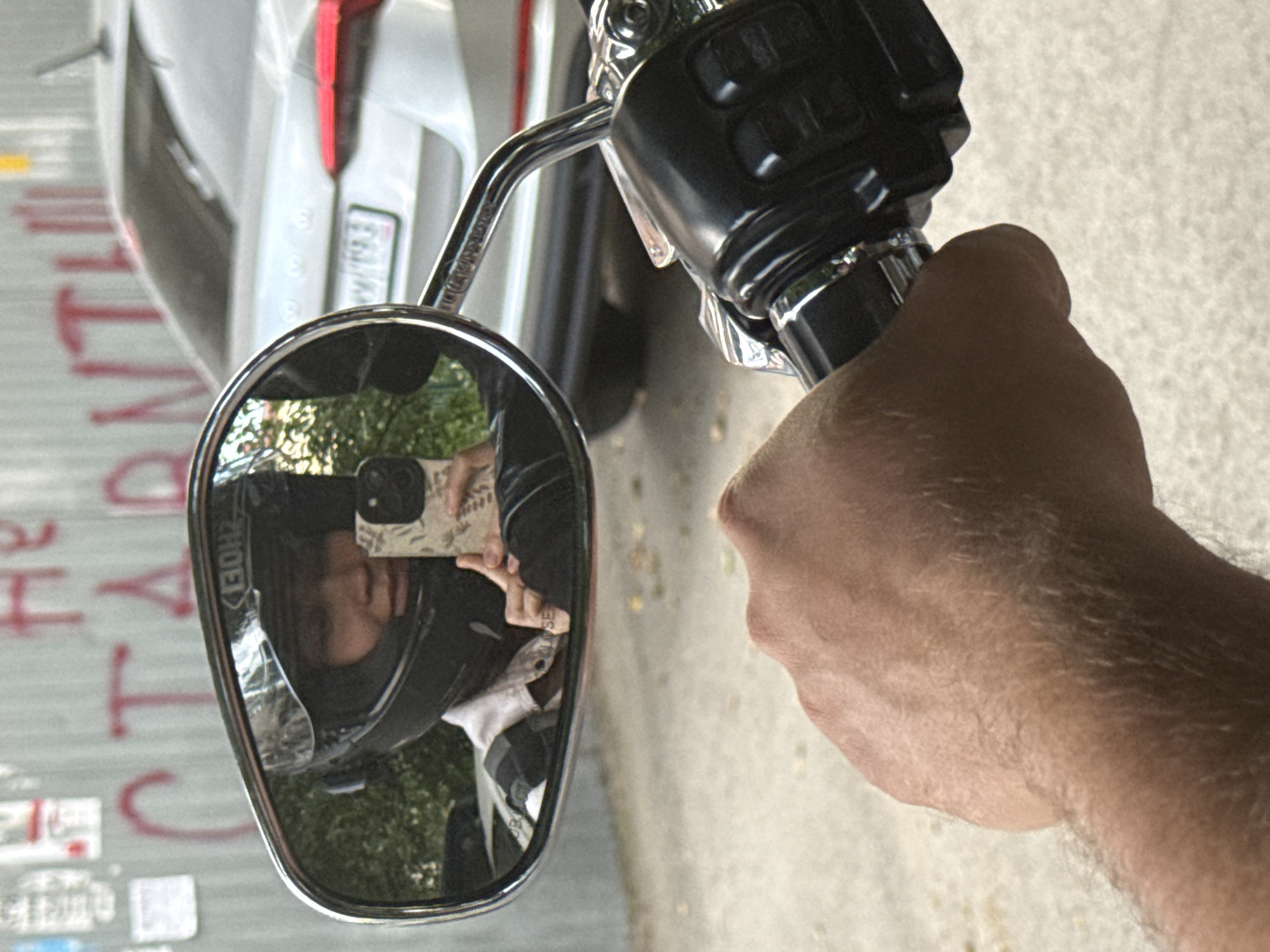 The image shows a close-up, angled shot of a person's right hand holding the handlebar of a motorcycle. The hand appears to be that of a Caucasian individual, with visible arm hair. The motorcycle handlebar is black and chrome, with a black grip and control buttons. A chrome rearview mirror is attached to the handlebar, reflecting a person wearing a black helmet and holding up a cell phone. The person in the mirror appears to be taking a selfie. The reflection also shows some greenery, possibly trees or bushes, behind the person.
In the background, a portion of a white vehicle is visible, along with a textured surface that appears to be a concrete or asphalt ground. To the left of the motorcycle and mirror, there is a vertical surface with red graffiti. The graffiti includes large, red, blocky letters, some of which are partially visible. Some smaller, white rectangular shapes are also present on this surface, possibly stickers or tags. The overall lighting suggests it is daytime, with no obvious indications of weather conditions. The scene is likely outdoors, given the presence of greenery and the vehicle. The person's activity of taking a selfie while holding the motorcycle's handlebar suggests they are either stopped or posing.