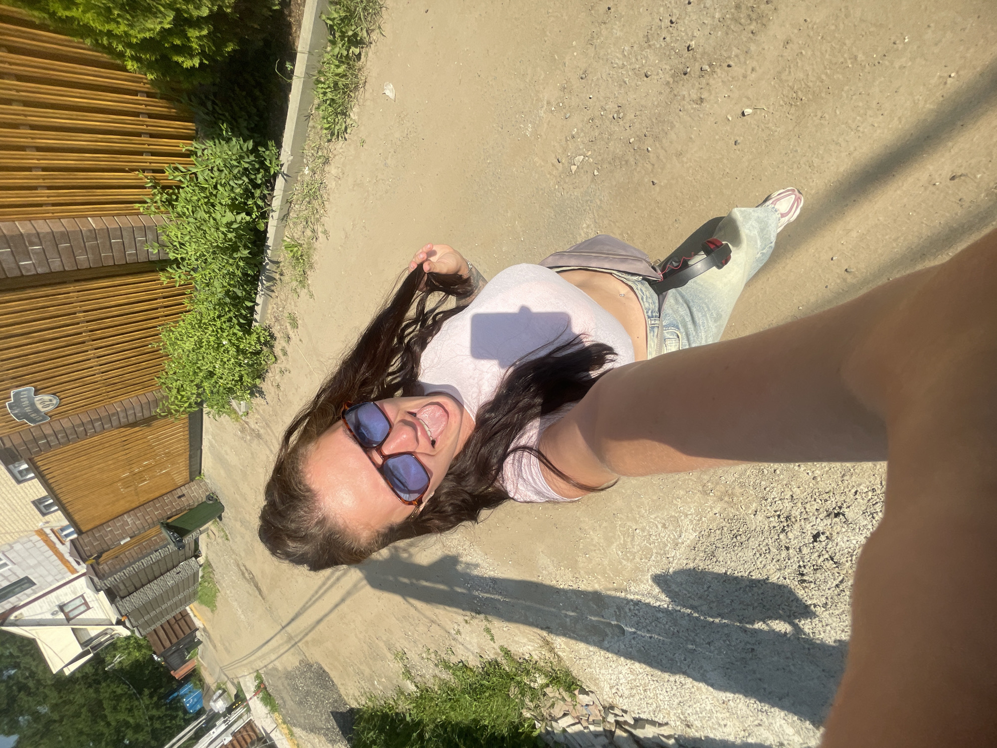 An outdoor, eye-level, medium selfie depicts a woman standing on a dirt road in an urban or suburban area. The woman has long, dark brown hair, wears oversized sunglasses, a light pink crop top, and light-wash jeans. She is sticking her tongue out and holding her hair with her left hand. A grey fanny pack is slung across her body. A long shadow of the woman is cast on the ground to her left, indicating the sun is behind her. The background shows a fence with vertical wooden planks, brick pillars, a green trash can, and houses with light-colored facades. There are also green bushes and trees visible. The overall environment suggests a residential street or alleyway within the city of Mosmetallokonstruktsiya Ont, Russia.