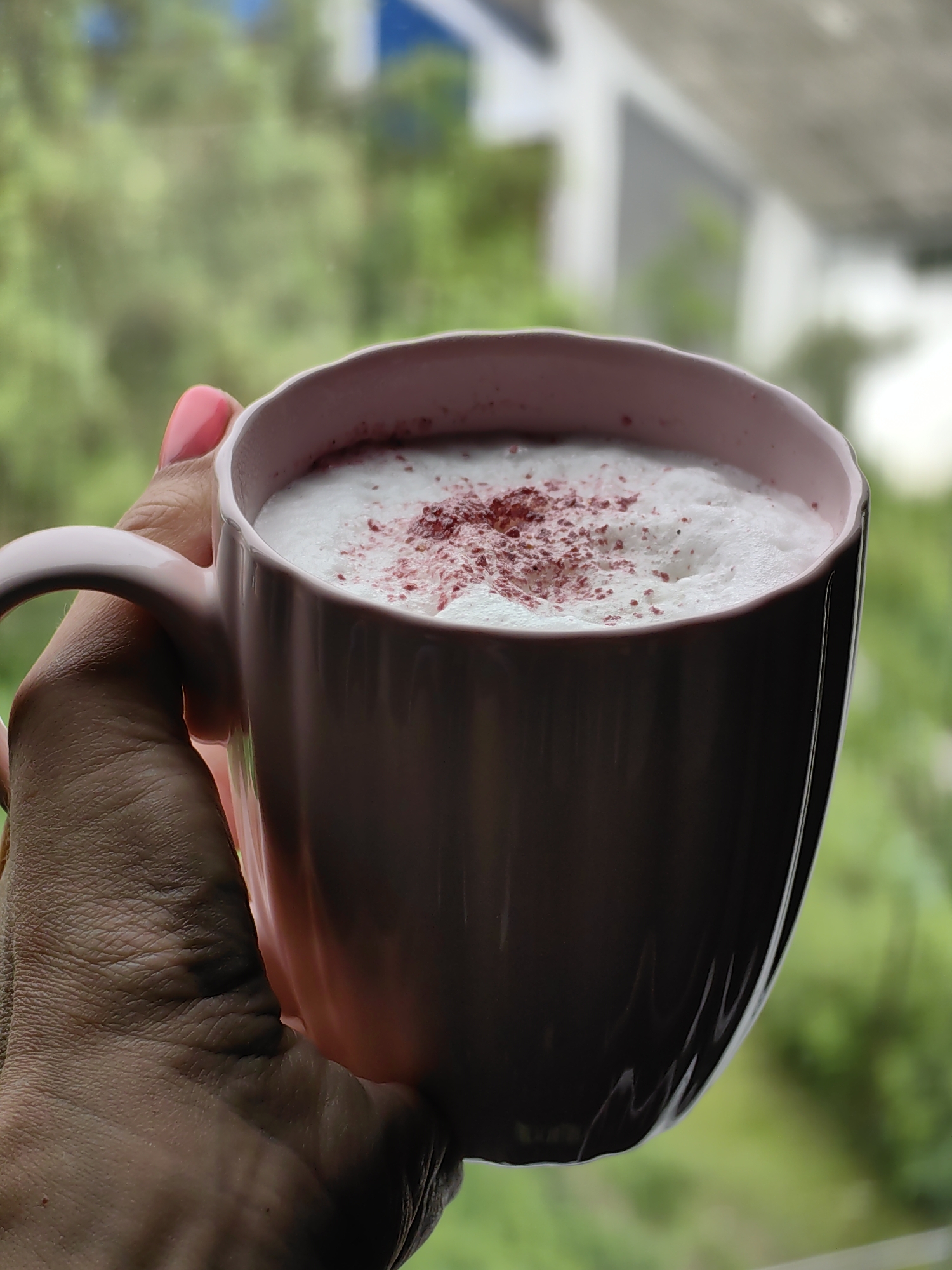 A person's hand with bright pink nail polish is holding a light pink mug filled with a frothy, white beverage, possibly coffee or tea. The drink is topped with a sprinkle of reddish-purple powder. The background is blurred, showing greenery and hints of white and blue structures, suggesting an outdoor or window setting. The image focuses on the mug and hand.