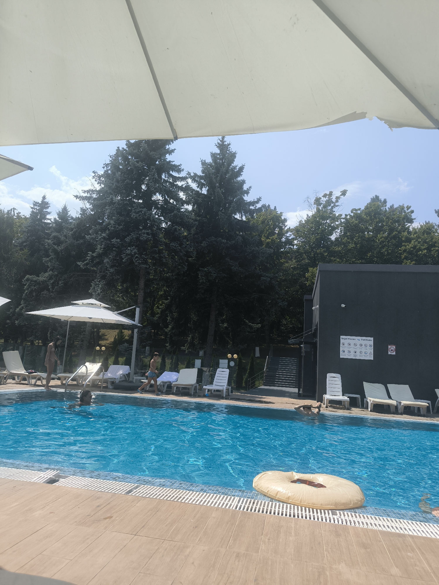 The image shows a sunny day at an outdoor swimming pool. In the foreground, a wooden deck leads to the bright blue water of the pool. Several lounge chairs are visible around the pool, and a large white umbrella provides shade. People are in and around the pool, with some swimming and others relaxing on the chairs. In the background, tall green trees form a natural border, and a modern building with a dark facade stands to the right, featuring a sign with various pool rules displayed on it. A inflatable ring floats in the pool. The scene suggests a recreational setting, likely a hotel or public pool, in Ustia, Moldova.
