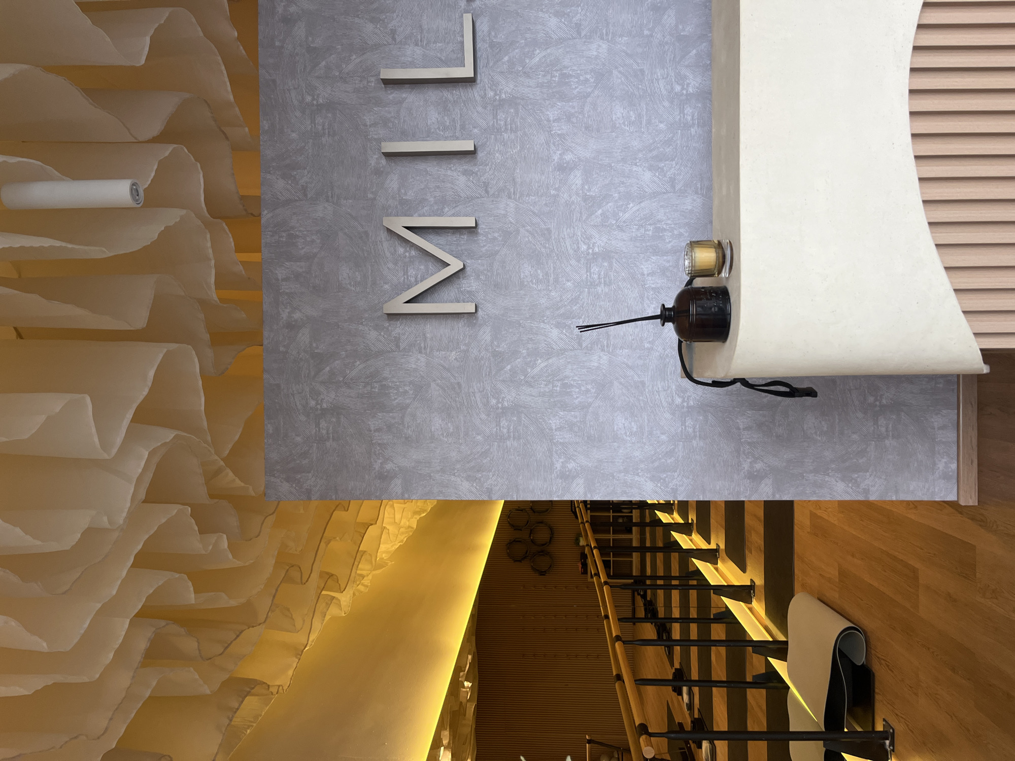 This media file depicts an elegantly designed interior space, likely a commercial establishment such as a boutique, spa, or hotel lobby, situated in San Bernardino Tlaxcalancingo, Mexico.
The upper section of the image showcases a light grey wall with a subtle, textured pattern of concentric circles. Mounted centrally on this wall are three large, three-dimensional metallic letters spelling "MIL". To the right of these letters, a smooth, curved white architectural feature, possibly a counter or decorative element, holds a dark brown diffuser bottle with reeds and a small glass container with a golden substance, likely a candle or oil. Behind this feature, a section of light-toned wooden slatted paneling is visible.
To the left, a striking ceiling or canopy is constructed from numerous layers of translucent, light-colored fabric or paper, creating a soft, undulating texture. A white cylindrical light fixture is nestled within these folds, casting a gentle glow.
The lower portion of the image reveals a modern staircase with wooden treads and dark metal risers or supports. Warm, indirect lighting illuminates the stairs and the adjacent wall, which features circular metallic decorative accents. A light-colored mat, possibly a yoga mat, is rolled and placed on one of the wooden steps. The flooring surrounding the staircase is made of light wood.
The scene is devoid of people, indicating no specific activity or event taking place at the moment the photograph was captured. The interior lighting creates an ambient atmosphere, without providing cues about the time of day or external weather.