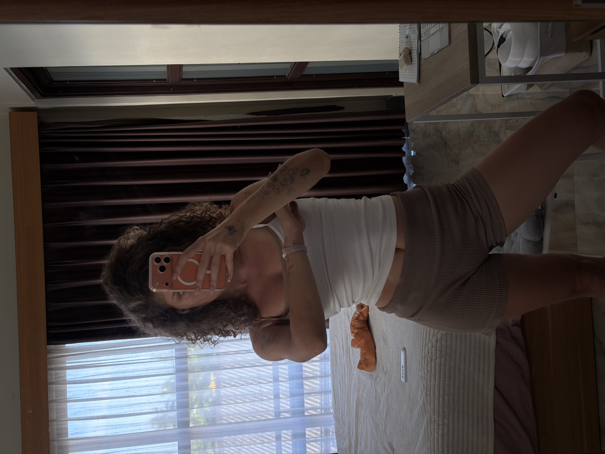 The image shows a woman taking a mirror selfie in a room. She is standing and wearing a white tank top and brown ribbed shorts. She has curly brown hair and several tattoos on her arms. In the background, there is a bed with an orange object on it and a window with sheer curtains. To the right, there is a desk with a laptop and some papers. The room appears to be a hotel room or apartment in North Kuta, Indonesia, given the presence of dark brown curtains and a white tiled floor, typical of tropical accommodations.