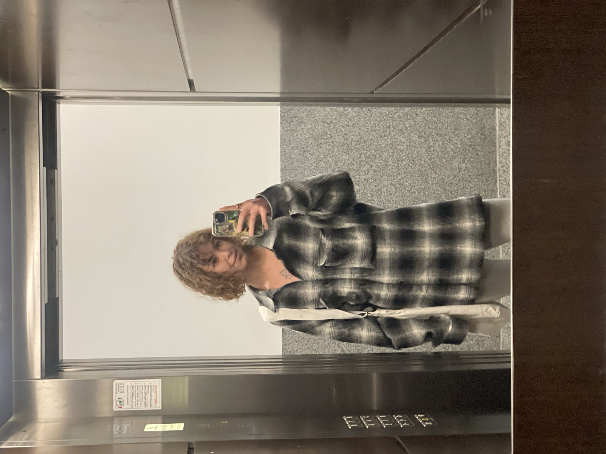 A person is taking a selfie in an elevator. The elevator's interior consists of light grey and white walls with stainless steel details, including the control panel. The person is wearing a grey and black plaid shirt and has curly blonde hair. The location is not definitively identifiable as Boreti, Montenegro based on the image.