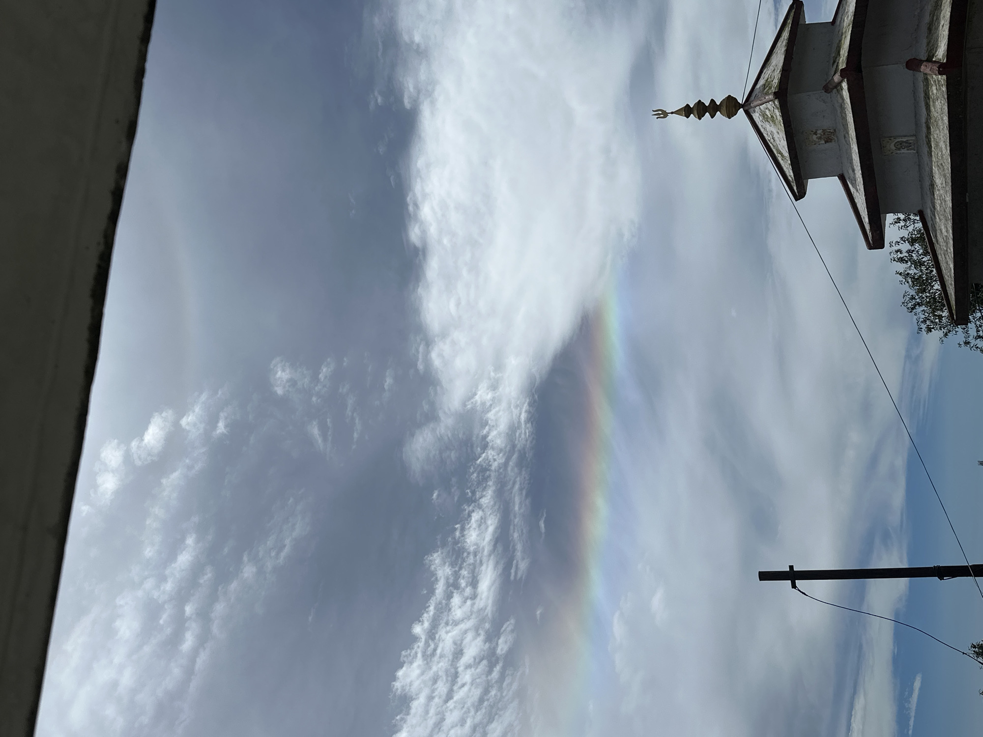 A wide-angle, eye-level shot captures a segment of the sky and the upper portion of a building in Haini, India. The sky is filled with a mix of light and dark gray clouds, with a faint rainbow visible in the middle left section of the frame. A dark, weathered pole stands to the left, with a wire attached to it, running towards the right and across the frame. On the right side of the image, the layered roof of a building is visible, topped with a gold spire. The building appears to be light-colored with red trim and decorative panels. The overall scene is overcast and atmospheric.