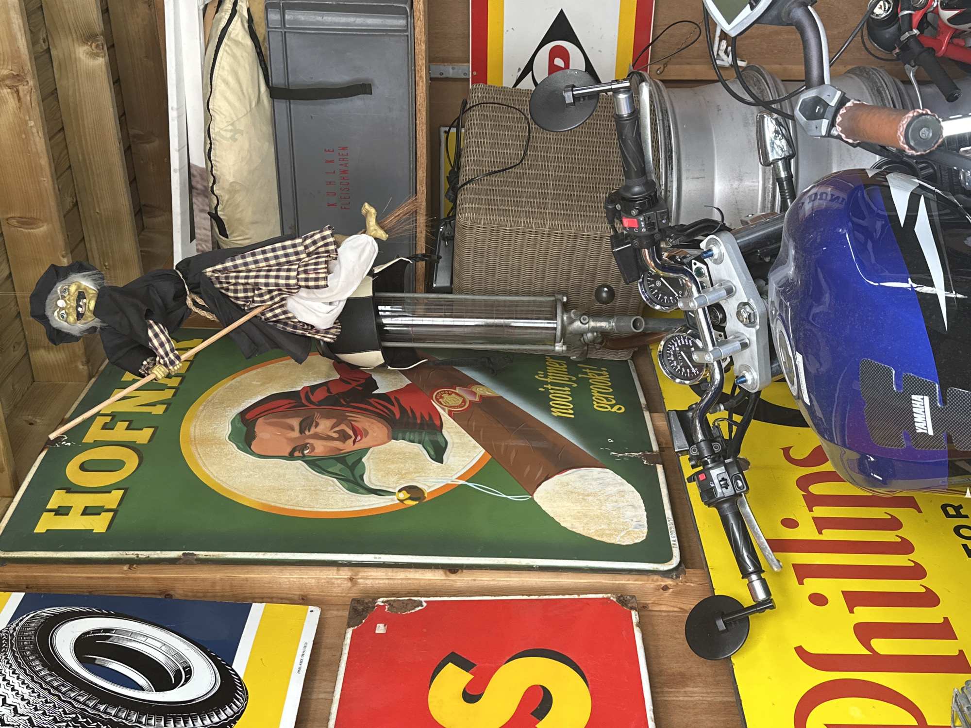 This is a photograph of a cluttered garage or storage area, possibly in Bruges, Belgium, containing various objects including vintage signage, a motorcycle, and a decorative witch figure.
The background features wooden planks forming the ceiling. On the left, a circular tire advertisement with a black and white tire on a blue and yellow background is visible. To its right, a large, green, rectangular sign with yellow lettering reads "HOFN". Below this, a circular image depicts a man's face with a green jester's hat and a wink, and a large brown cigar. The text "nooit fijner gerookt!" is also present on this sign.
A decorative witch figure, dressed in black with a plaid skirt and white apron, hangs from the ceiling, holding a broomstick. Behind the witch, on a shelf, is a grey plastic storage container with German text that reads "KUHLKE FLEISCHWAREN". Below this are wicker baskets.
In the foreground, a blue motorcycle is parked. The handlebars, instrument panel with dials, and a portion of the fuel tank are visible. The motorcycle is positioned in front of a yellow sign with red lettering that appears to read "Phillips". Additional motorcycle parts and accessories are scattered around. Another red sign with a black and yellow design is partially visible on the right. The overall impression is one of a collection of items, possibly related to automotive history or vintage advertising, displayed in a disorganized manner.