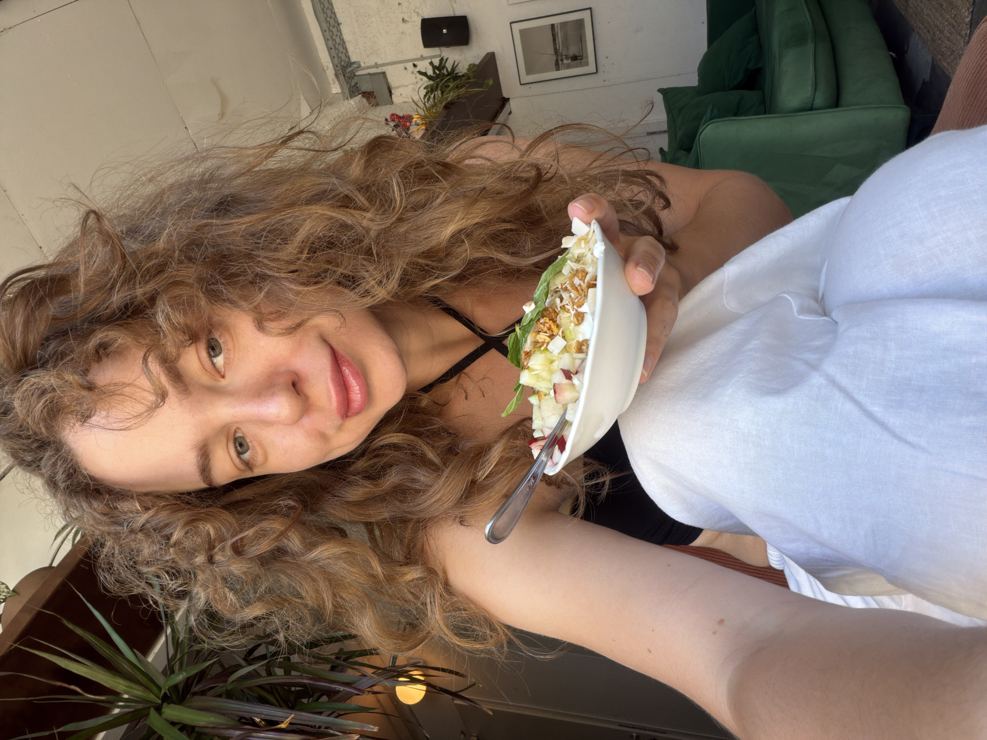 A woman with curly brown hair takes a selfie while holding a bowl of food. She is seated, wearing a black top and white pants. The background shows a green sofa, a plant, and a framed picture on the wall, suggesting an indoor setting possibly in Tel-Aviv, Israel.