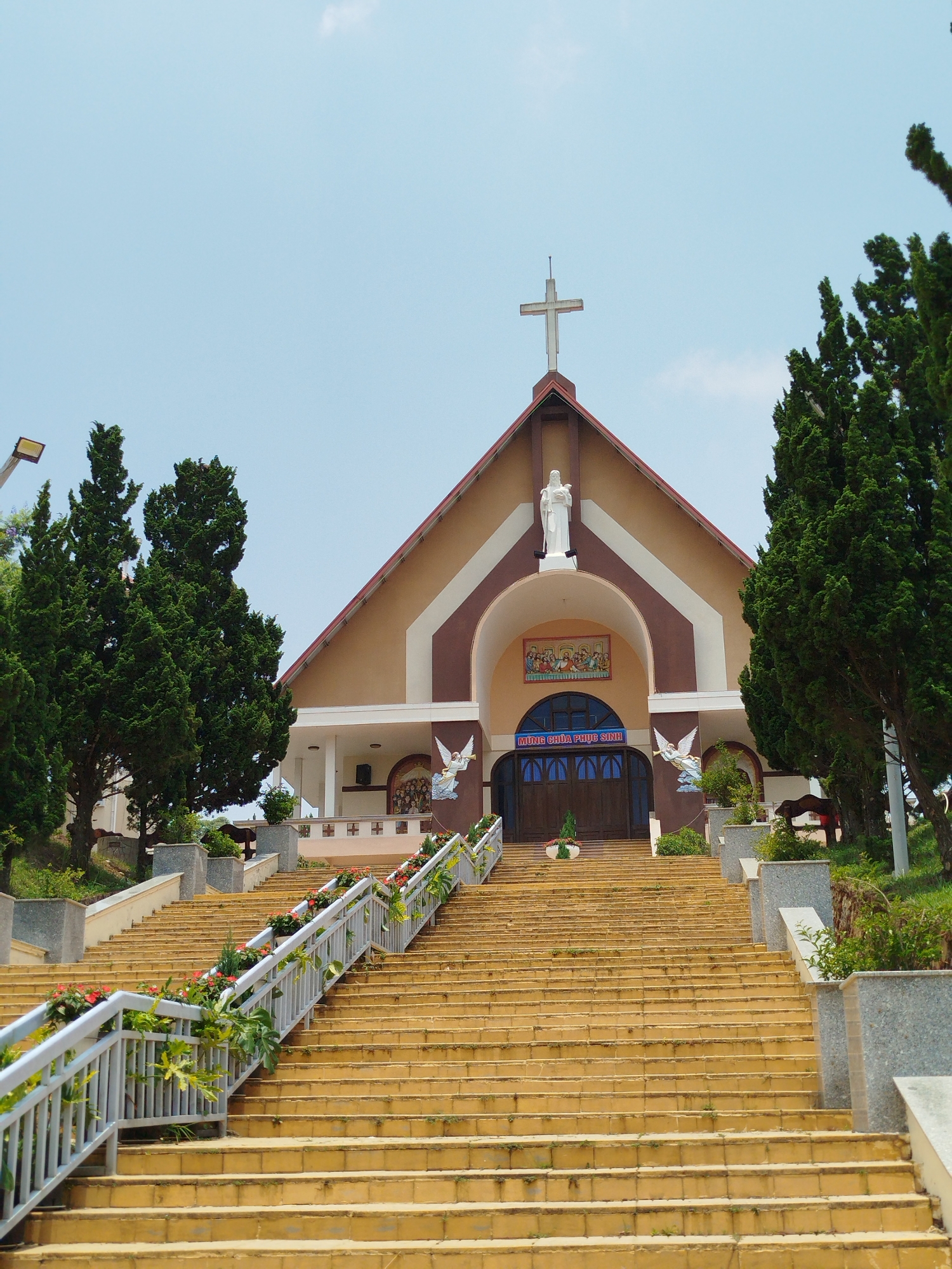 The image depicts the exterior of a church, located on a hill in Lang Biang - Da Lat, Vietnam. A long staircase leads up to the main entrance of the building. The church has a beige and brown facade, with a peaked roof topped by a cross. A white statue stands above the entrance archway. Two angel figures are depicted on either side of the entrance. Trees flank the church on both sides. The staircase is decorated with flowers along the railings. The sky above is light blue.