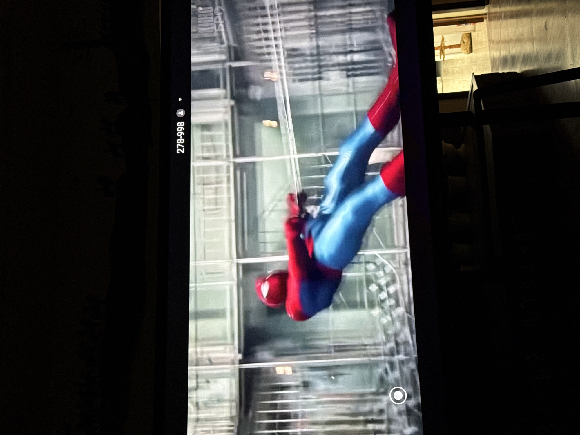 The image shows a television screen displaying a scene from a movie or TV show. The main subject is a person in a red and blue superhero costume, appearing to be Spider-Man, in a dynamic pose against a blurred background of a building with glass windows and metal railings. The on-screen display includes a timestamp "278-998" and a logo that appears to be "КИНО ЕРО". The image is taken in a dimly lit room, with the lower part of the frame showing a dark table or furniture. There is no indication of the location being in Nyurba, Russia, and this detail cannot be inferred from the visual information provided.