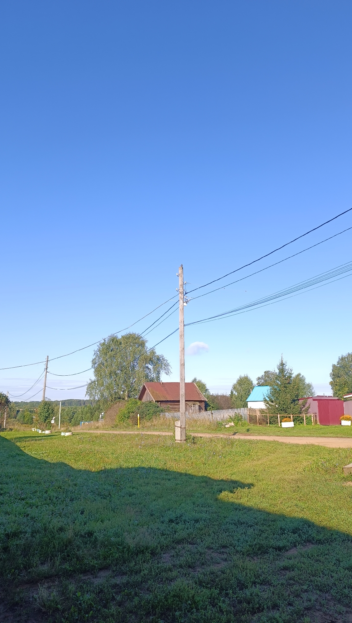 The image depicts a rural landscape under a clear blue sky. In the foreground, a well-maintained grassy area casts a long shadow, indicating it is likely morning or late afternoon. A dirt road runs horizontally across the mid-ground.
Several buildings are visible: a rustic wooden house with a reddish-brown roof, a white building with a bright blue roof, and a red shed. These structures appear to be part of a village or rural settlement. A wooden utility pole with power lines stands prominently in the center of the frame, with another similar pole visible to the left. Trees, including a large deciduous tree with sparse foliage and several evergreen trees, are scattered throughout the scene, with a dense treeline forming the background. A wooden fence separates the road from some of the properties. There are also white objects in the distance, which may be beehives or small structures.
The scene conveys a peaceful, sunlit rural environment. There are no people or animals visible in the image. The overall impression is one of a quiet, residential area in a village setting. The prompt specifically asks for the location in the city of Karsha, Russia, however, the image itself does not contain any identifying markers or context to confirm this location. This description is solely based on the visual elements present in the photograph.