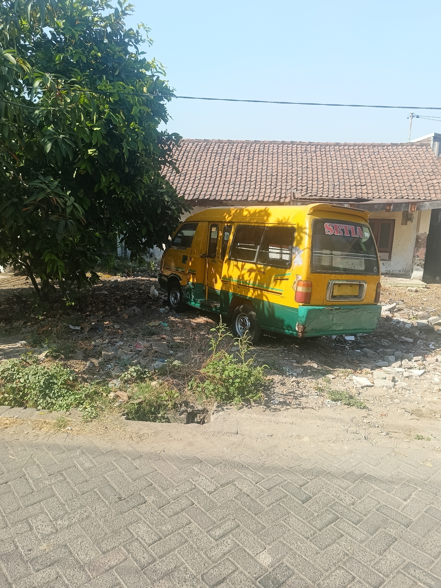 A yellow and green van is parked on a dirt lot next to a brick building with a tiled roof. A large tree with green leaves is to the left of the van, casting a shadow over it. The van has the word "SETIA" written in red on the back window. The lot is filled with debris, including rocks, trash, and overgrown weeds. The ground in the foreground is paved with interlocking bricks. The sky is clear and blue. The scene appears to be in a rural or semi-rural area.
