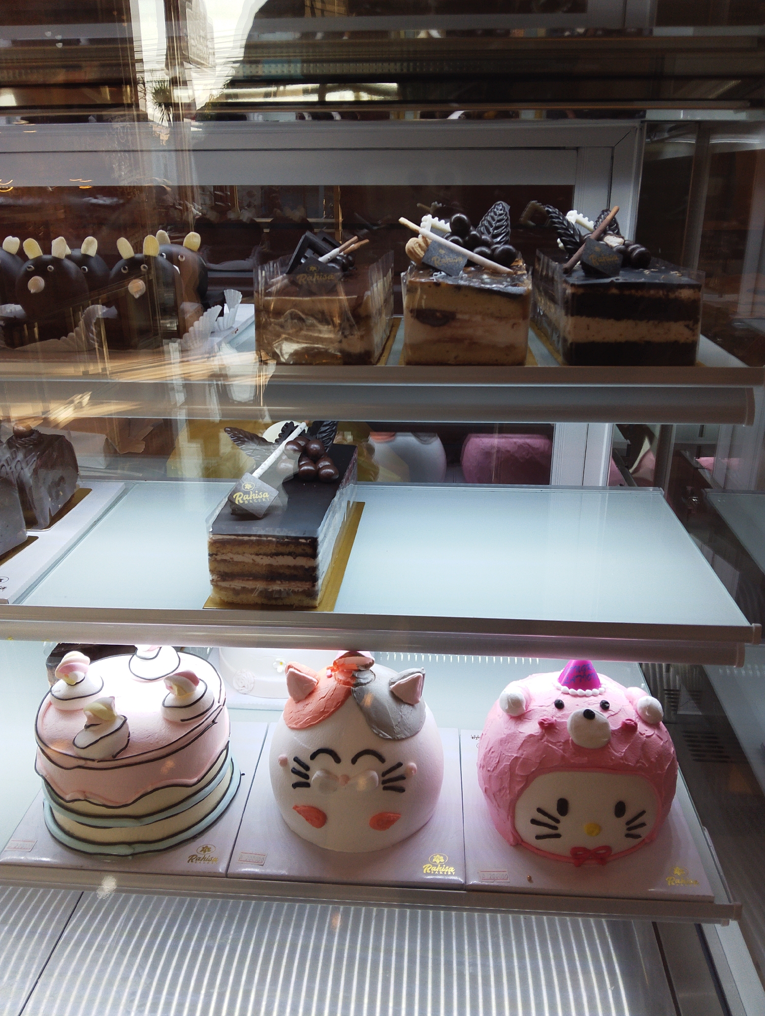 In Manaruwi, Indonesia, a bakery display case exhibits an assortment of cakes. The topmost shelf features several chocolate cakes, including two rectangular slices adorned with chocolate decorations and a whole cake partially visible. Below this, a rectangular chocolate cake with layered cream and chocolate frosting sits on the middle shelf, alongside other desserts and pink-colored pastries. The bottom shelf showcases three distinct cakes: a pastel-colored layered cake decorated with marshmallow-like toppings, a white cake shaped like a cat's head with orange ears and black facial features, and a pink cake resembling a character, possibly Hello Kitty, with a party hat and bow. The display case is made of glass, reflecting the surroundings.