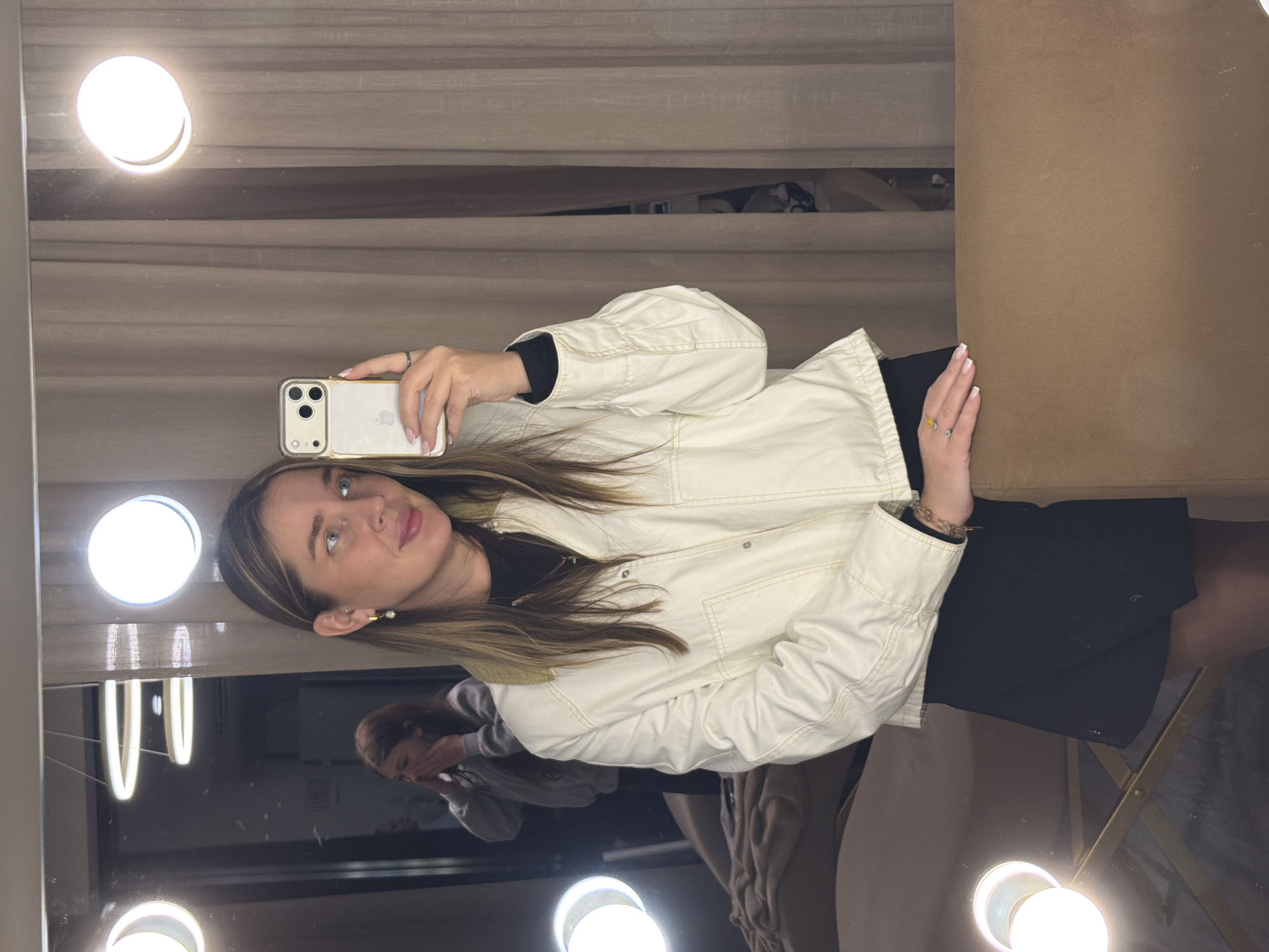 A woman with blonde hair is taking a selfie in a mirror. She is wearing a white jacket and a black dress. The mirror has lights around it. There is another person visible in the reflection, standing behind her. The scene is indoors. The location is Moscow, Russia.