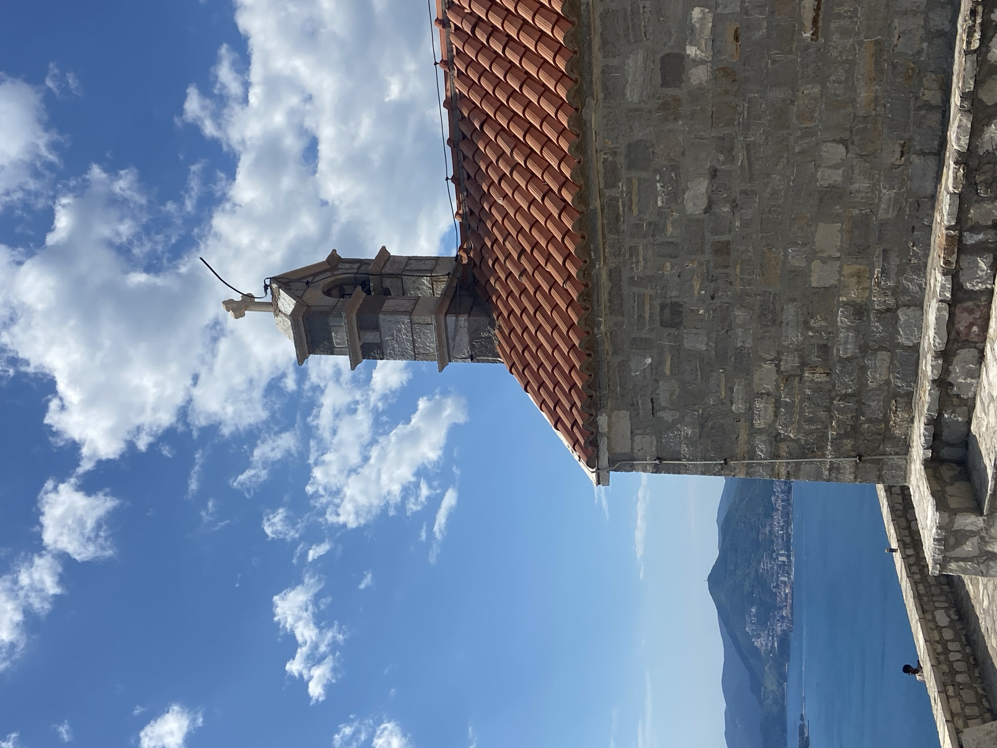 The image presents a scenic coastal landscape, rotated 90 degrees counter-clockwise, featuring historic architecture in the foreground against a backdrop of sea and mountains under a bright sky.
In the left half of the frame, a robust stone building, likely a chapel or part of a larger complex, stands prominently. Its walls are constructed from irregular grey stone blocks, and it is topped with a distinctive red-orange terracotta tiled roof. A small, elegant stone bell tower, or campanile, rises from the roofline, featuring an open belfry and a simple cross at its very top.
Below the building and extending across the middle and right side of the image is a wide expanse of deep blue water, indicative of a sea or bay. On the distant horizon, a range of verdant mountains slopes down to meet the water, with a town or settlement visible along the coastline at their base. To the far right, a stone wall or parapet, similar in construction to the main building, frames the view, and a small, partially visible person is seen leaning over it, observing the distant panorama.
The sky above is a brilliant, clear blue, dotted with scattered white cumulus clouds. The strong, direct sunlight suggests the time of day is likely midday or early afternoon, contributing to the vibrant and well-lit scene. The architecture and coastal features strongly evoke the known characteristics of Sveti Stefan, Montenegro, renowned for its picturesque island-village and Mediterranean setting. There is no visible text in the image.