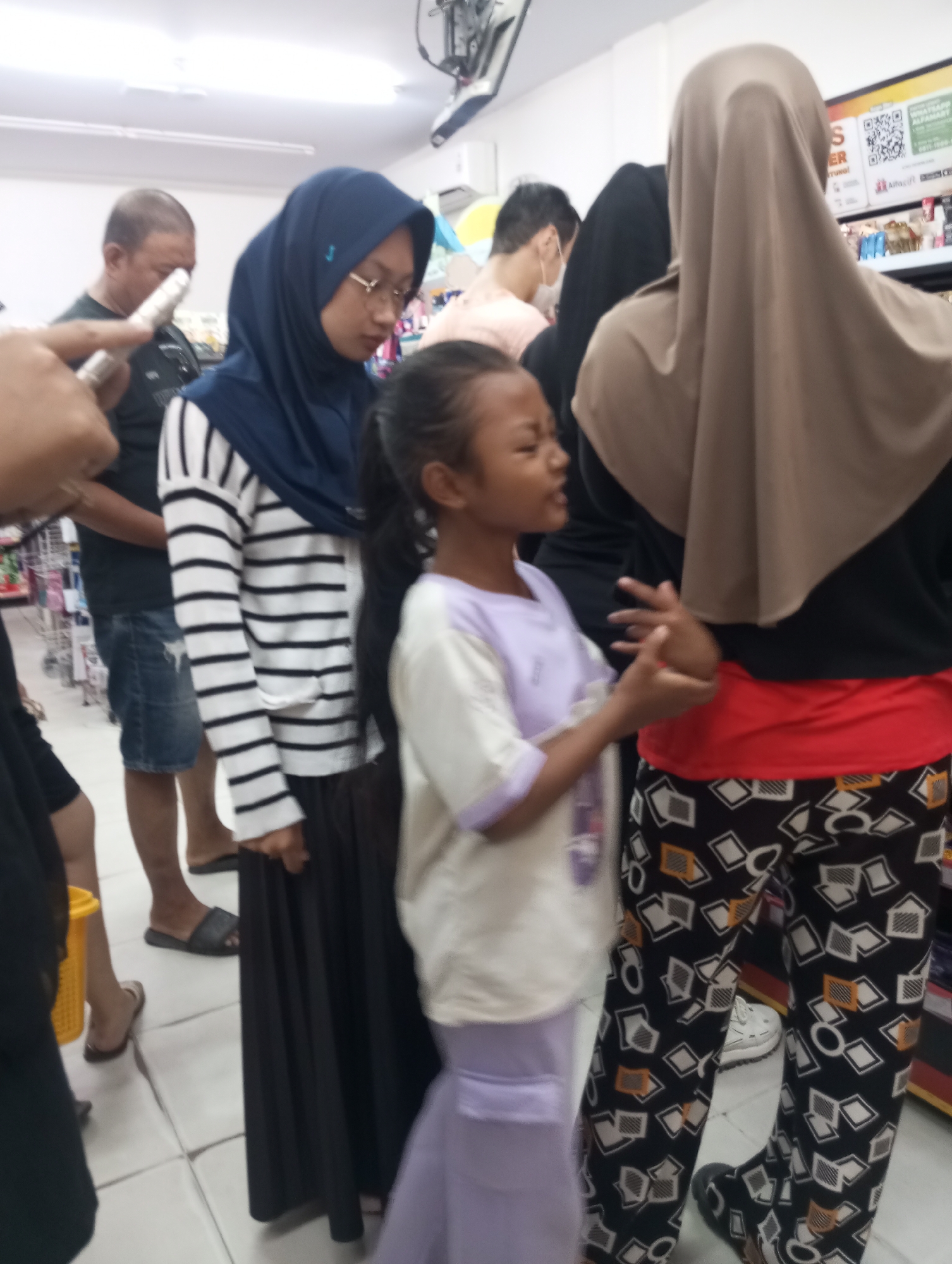 The image depicts a scene inside a retail store, likely a convenience store, in Tanjung Gading, Indonesia. Several individuals are visible, including a young girl in the foreground wearing a light purple shirt and skirt, her right hand gesturing outwards. Behind her, a woman in a blue hijab and glasses wears a striped long-sleeved shirt and a black skirt. To her right, another woman is seen from the back, wearing a beige hijab, a black top, and red fabric around her waist, with patterned black pants. A man in a dark t-shirt and denim shorts is positioned to the left of the woman in the blue hijab. The environment includes shelves stocked with products, a ceiling with lighting, and a tiled floor. A yellow basket is visible on the floor. The overall impression is that of a busy shopping environment with people in a queue or browsing.