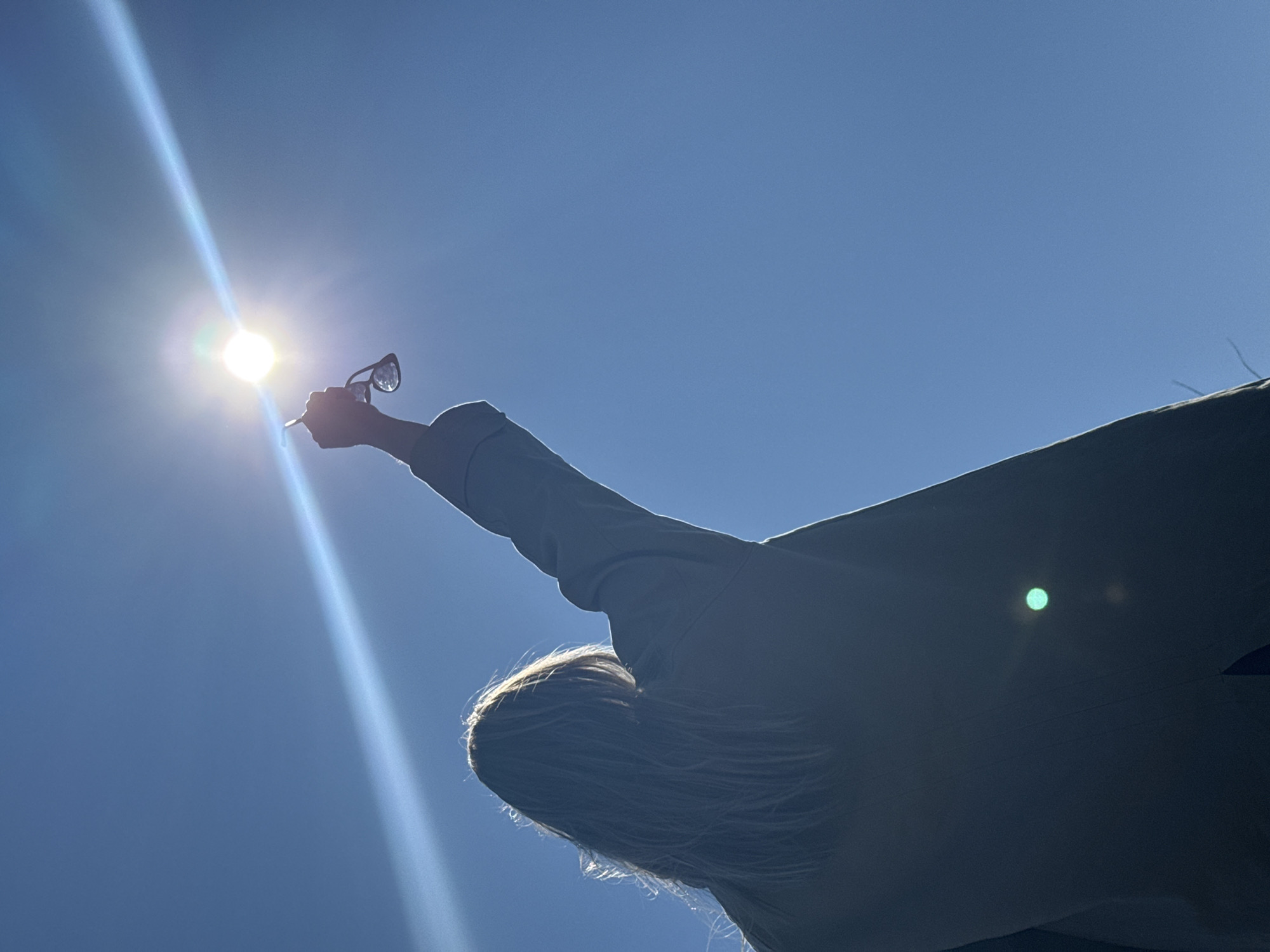 A low-angle shot shows a person holding eyeglasses toward the sun in a clear blue sky. The sun is high and bright, creating a lens flare. The person is silhouetted, with their blond hair visible. They are wearing a light-colored jacket or coat. The scene appears to be outdoors on a sunny day. The location is not discernible from the visual information.