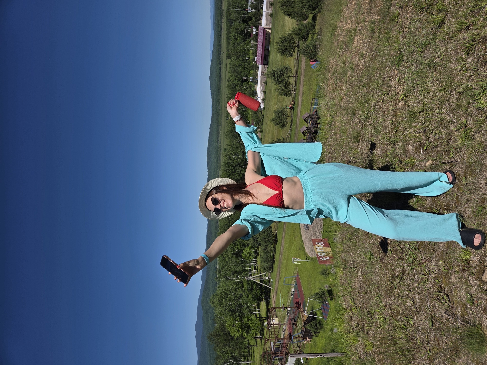 A woman stands on a grassy hill, holding a smartphone in her raised right hand and a red tumbler in her raised left hand. She wears a red bikini top, light blue wide-legged pants, a matching light blue open shirt, a straw hat, and sunglasses. The background displays a lush green landscape with trees, a playground, and distant hills under a clear blue sky. The setting appears to be a rural or park-like area.