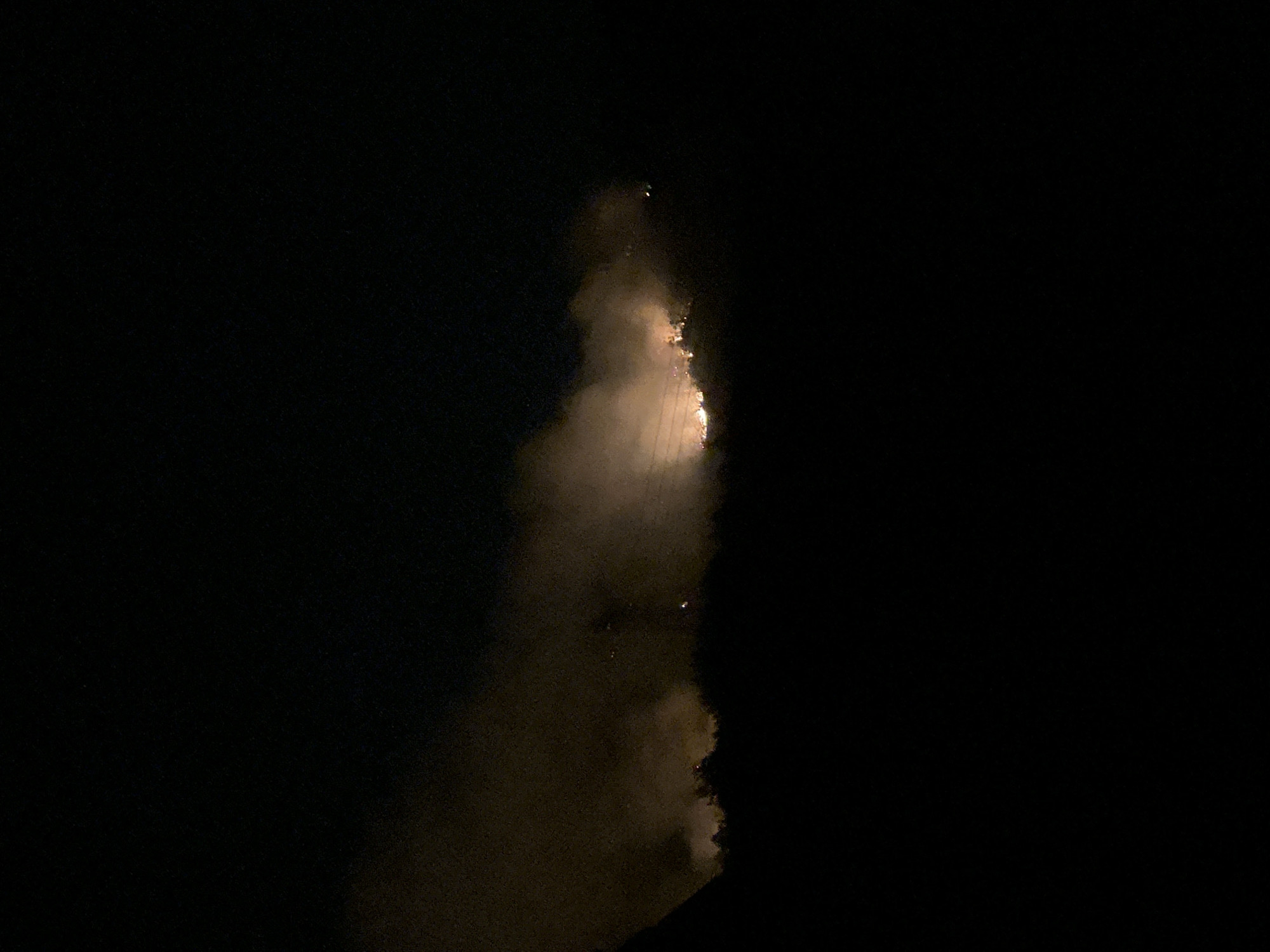 A nighttime photograph depicts a hillside ablaze with fire, casting an orange glow and smoke into the dark sky. The fire appears to be concentrated along a ridgeline, with flames visible amidst trees and undergrowth. Smoke billows upwards, partially obscuring the flames and creating a hazy atmosphere. The foreground is dominated by a dark silhouette of the land, suggesting a low vantage point for the photographer. The location is identified as Bijele Poljane, Montenegro.