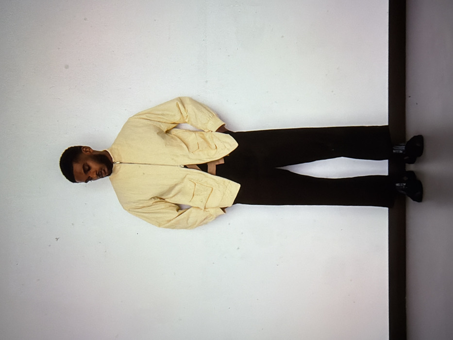 A full-body, frontal photograph depicts a solitary adult male standing against a plain, off-white wall. The man, with dark skin, short dark hair, and a trimmed beard, is looking downwards and slightly to his left. He wears a light yellow, textured, zip-up jacket with a stand collar and two flap pockets, partially unzipped to reveal a brown and black patterned garment underneath. He also wears black, wide-leg trousers that are cropped above the ankle, and black dress shoes. His hands are in his pockets. The floor is dark gray, meeting the wall at a dark horizontal line. The environment appears to be an indoor studio or minimalist setting. No visual information within the image identifies the location as being in the city of London, United Kingdom.