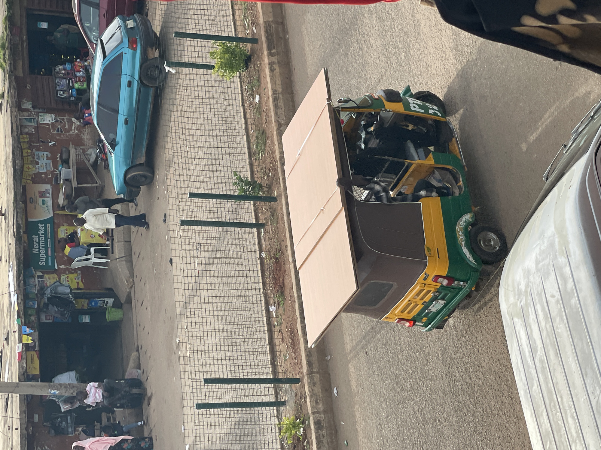 This is a medium-distance, high-angle shot of a street scene in a city. In the foreground, the roof of a white vehicle is visible, with horizontal indentations. A three-wheeled auto-rickshaw, yellow and green with a brown canopy, is positioned in the center of the frame, traveling away from the viewer. The auto-rickshaw has a large, flat wooden board strapped to its roof. There are people inside the auto-rickshaw, and the license plate appears to read "PT 32". The auto-rickshaw is on a paved road. Behind the auto-rickshaw, a metal chain-link fence runs horizontally across the middle ground. Beyond the fence, a row of shops and market stalls line the street. One shop has a sign that reads "Nerat Supermarket". Several people are visible near the shops, some standing, some sitting. A turquoise hatchback car is parked on the street in front of the shops, and the back of a red car is visible to the right. The background shows buildings and more market activity. The scene appears to be in a busy, commercial area.