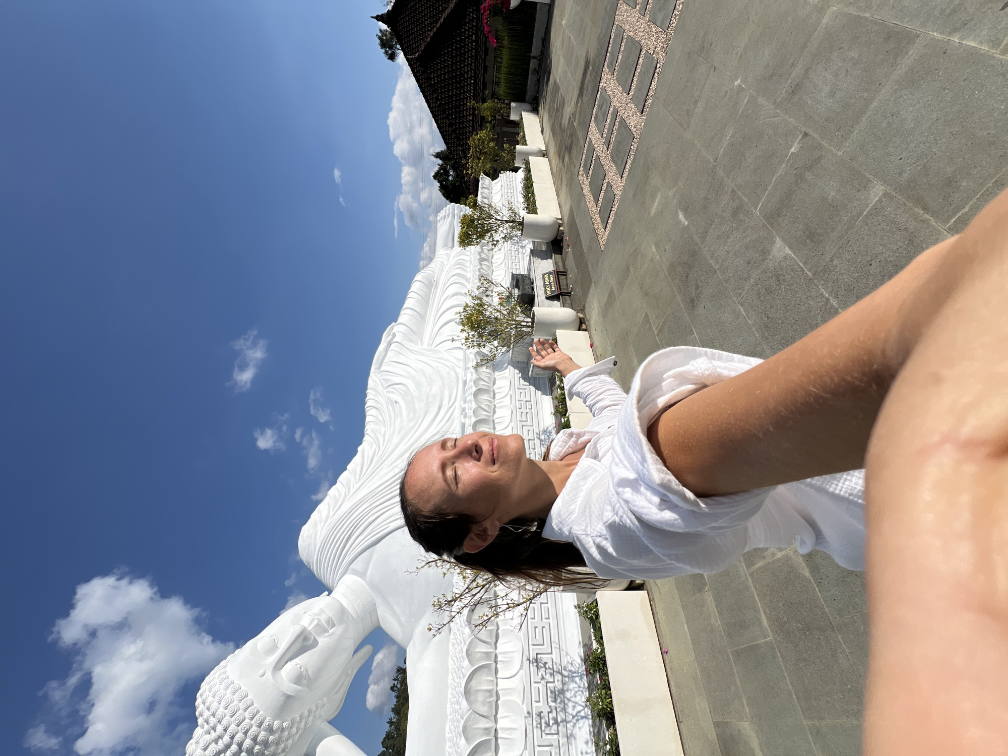 A selfie is taken by a woman standing in front of a large, white, reclining Buddha statue in Pupuan, Indonesia. The woman is facing away from the camera, with her eyes closed and arms outstretched, appearing to be in a meditative or welcoming pose. She is wearing a white shirt. The Buddha statue is detailed and positioned on a raised platform with decorative elements. The ground is paved with large, square stones, and there are potted plants and a building with a dark, tiled roof in the background. The sky is a clear blue with scattered white clouds, and the sun is shining, casting shadows.