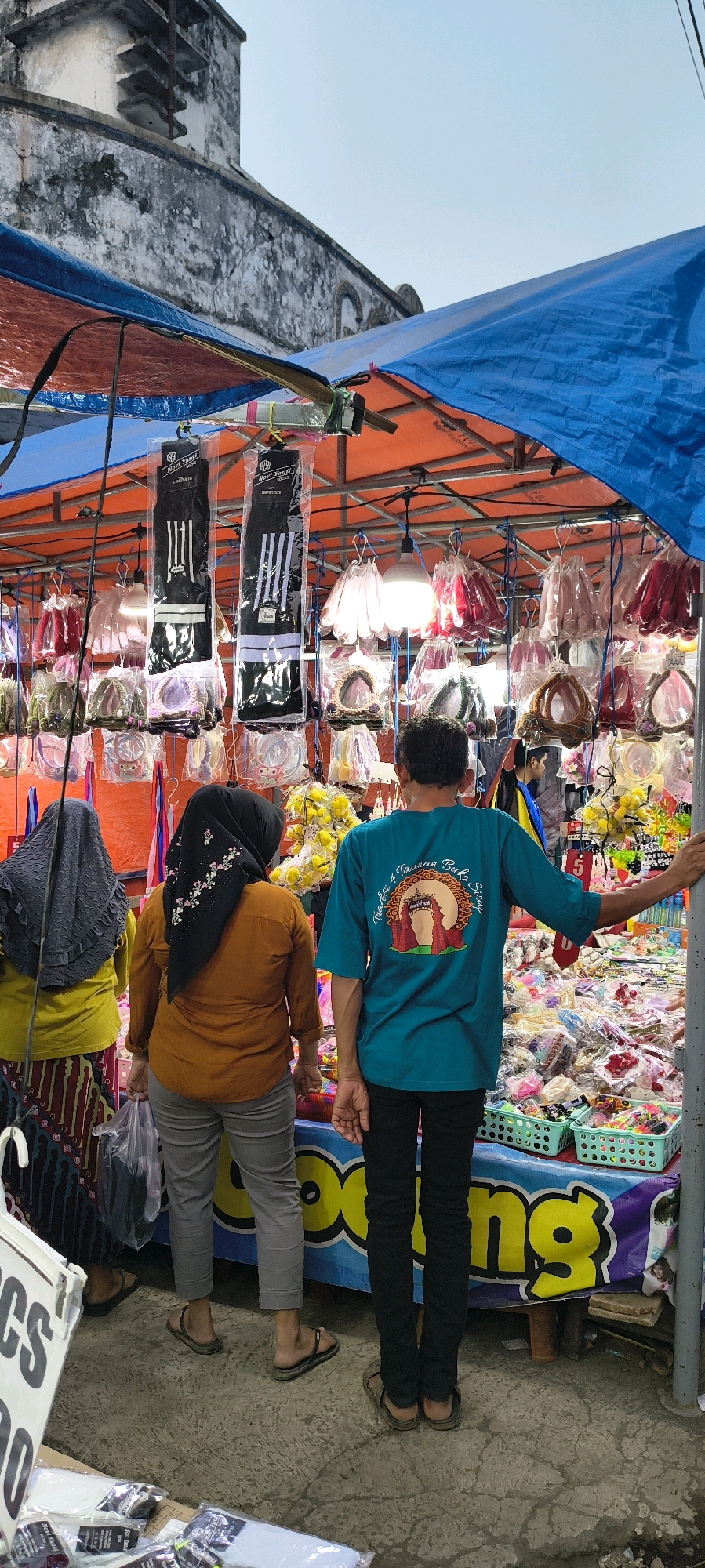 The image captures an outdoor stall in Trusmi Wetan, Indonesia, displaying various goods under a blue and orange tarpaulin canopy. Three individuals are visible. On the left, a person wearing a dark grey hijab and a yellow shirt stands partially visible. In the center, a person wearing a black hijab and an orange shirt is looking at the stall. On the right, a person in a teal t-shirt with a graphic design on the back and black pants stands with their right arm resting on a pole. The stall is laden with merchandise, including bags, accessories, and what appear to be socks packaged in clear plastic. The background shows an aged, weathered stone building. The ground is concrete with visible cracks. A large banner with yellow lettering on a blue background is draped across the front of the stall.