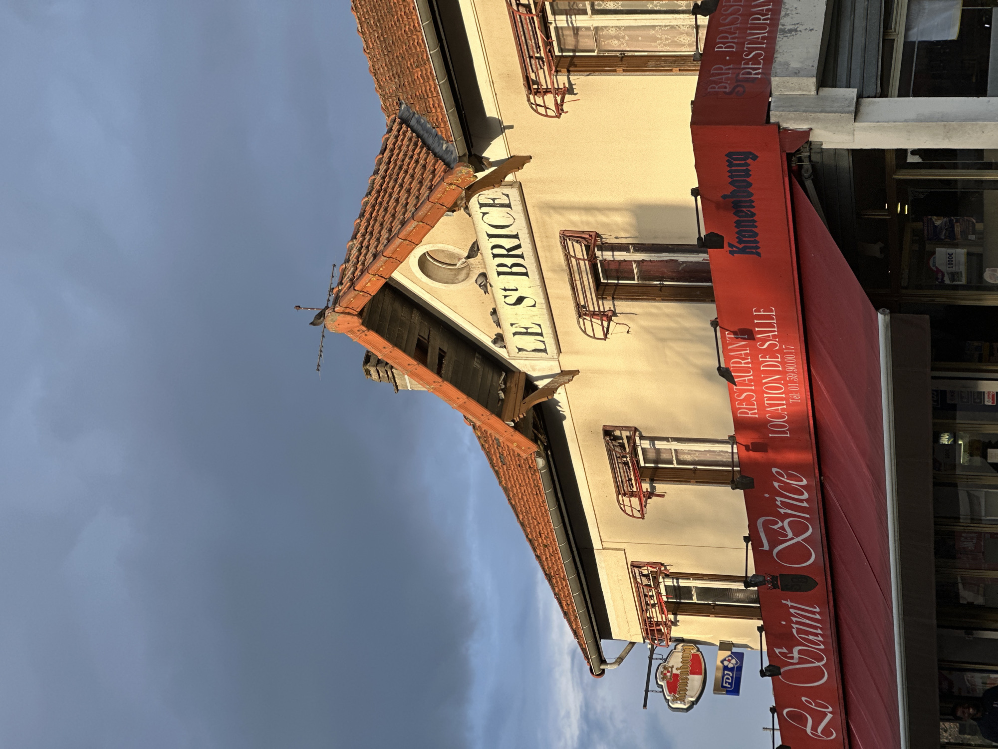 The image depicts the front of a building in Saint-Brice-sous-Forêt, France, identified as "Le Saint Brice," a restaurant and bar-brasserie. The building has a red-tiled roof and a light-colored façade with windows adorned with red metal balcony railings. A red awning stretches over the entrance, displaying the names "Le Saint Brice" and "Kronenbourg." There is a sign indicating "RESTAURANT LOCATION DE SALLE" with a telephone number. Additional signs include a "Kronenbourg" beer advertisement and an "FDJ" (Française des Jeux) sign. The sky above is overcast with dark clouds. There is evidence of additional storefronts to the right of this building.