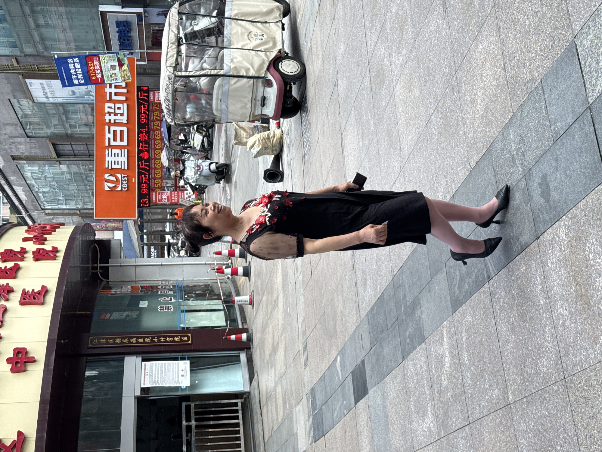 A middle-aged Asian woman with her hair pulled back in a bun is walking to the right on a paved sidewalk in Jiangjin, China. She is wearing a black dress with red floral embellishments on the neckline and sheer sleeves, and she has on dark high heels and pink hosiery. She is holding a dark-colored smartphone in her right hand. The woman is smiling as she walks.
In the background, there is a large orange sign with white Chinese characters that reads "重百超市" (Chongbai Supermarket). Below that, there are advertisements for various food items with prices. Further in the background, there are several motorized scooters and a small white and tan electric vehicle that resembles a rickshaw. To the left of the woman, there is a building with a dark awning and glass doors. A vertical sign on the building has Chinese characters on it. Red and white striped cones are placed along the edge of the sidewalk. The overall environment appears to be a commercial street in an urban area.