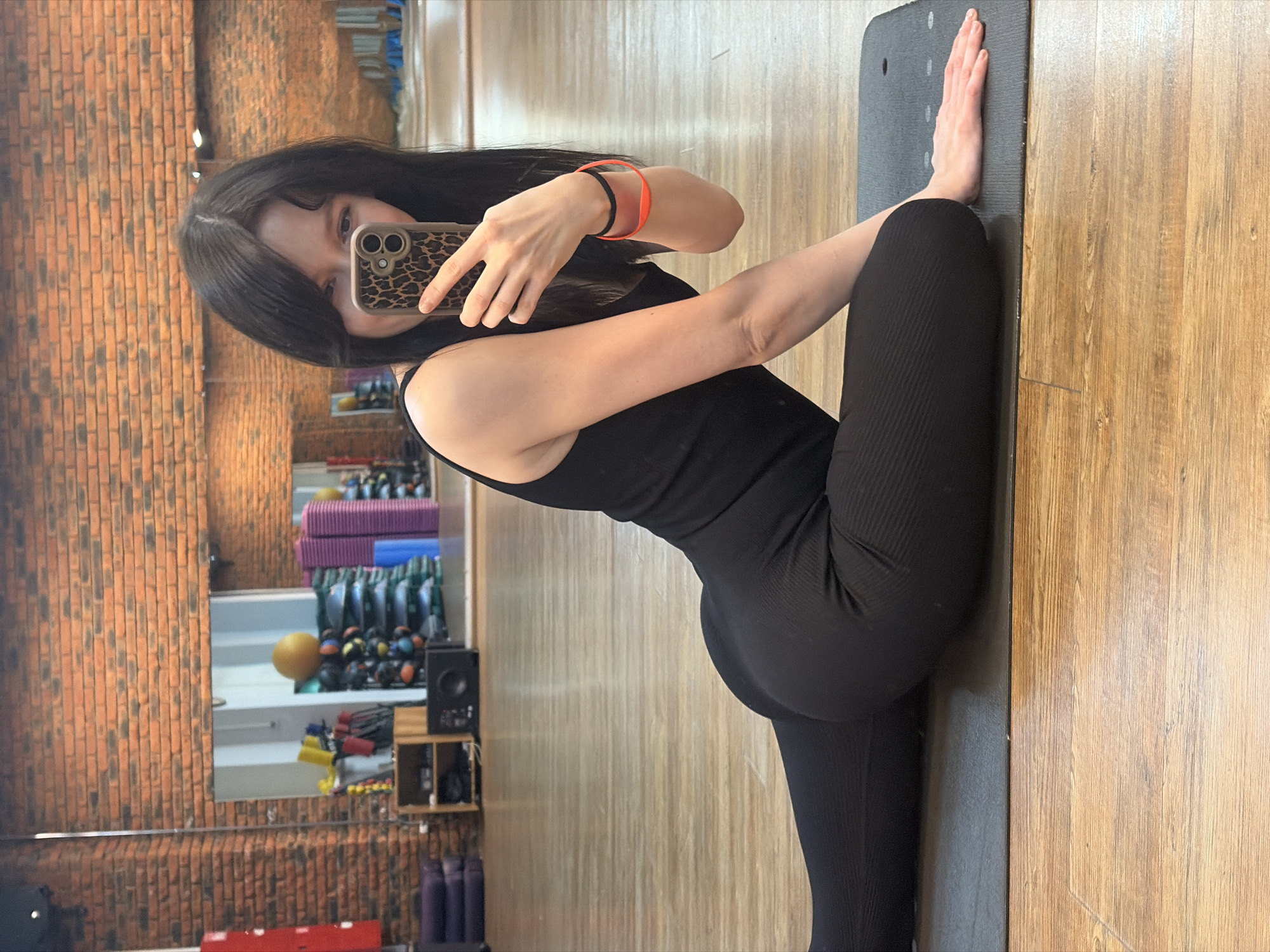 A woman is posing in a gym setting, taking a selfie with her mobile phone. She is wearing a black one-piece jumpsuit. She is kneeling on a black yoga mat. The background shows a gym with workout equipment, weights, and yoga mats. The image is likely taken in Chaikovsky, Russia, based on the location information provided.