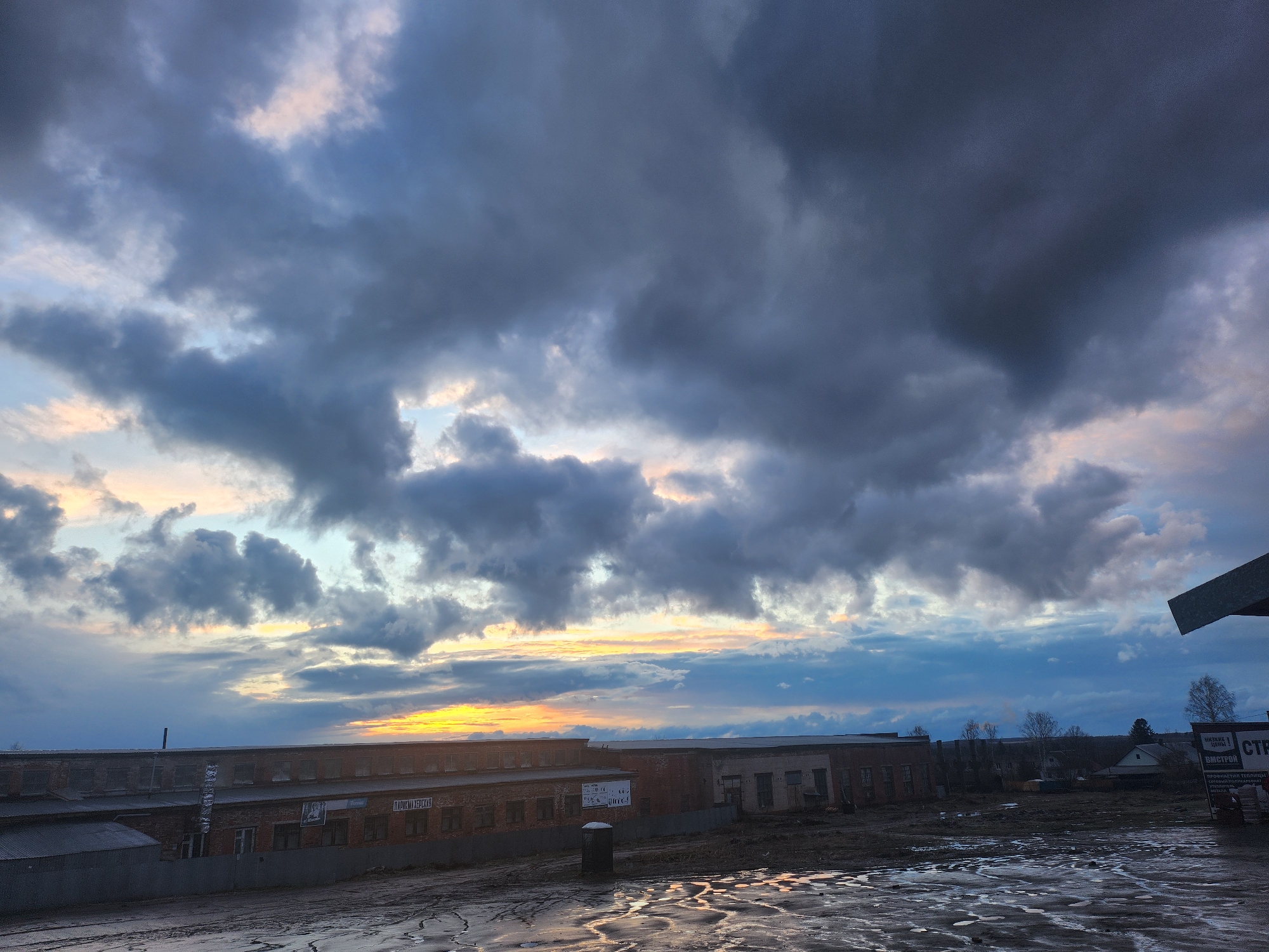 This image captures a semi-industrial landscape in Krasnogorskoye, Russia, under a dramatic, heavily clouded sky during either sunrise or sunset. The sky is dominated by thick, dark grey clouds, with vibrant bands of orange, yellow, and pink light illuminating the horizon, suggesting the sun is low.
The foreground features a wet, uneven ground with numerous puddles reflecting the sky, indicating recent rain or snowmelt. In the mid-ground, several low-rise, aged brick buildings, resembling factories or warehouses, extend across the frame. One building on the left has a tall ladder propped against it and displays Cyrillic signs, including "ПАРИКМАХЕРСКАЯ" (Hairdresser's), "АМЕРИКАНСКАЯ" (American), and "Уютная Уборка" (Cozy Cleaning). Further to the right, another building stands, with a portable toilet in front of it.
On the far right, a large sign or billboard is partially visible, featuring text such as "НИЗКИЕ ЦЕНЫ!" (LOW PRICES!), "ВМСТРОЙ" (VMSTROY, likely a company name), and mentions of construction materials like "ПРОФНАСТИЛ, ТЕПЛИЦЫ" (profiled sheeting, greenhouses). Beyond the buildings, bare trees and hints of other distant structures are visible. No people are present in the scene. The overall mood is quiet and slightly stark, with the dramatic sky providing a striking contrast to the industrial setting.