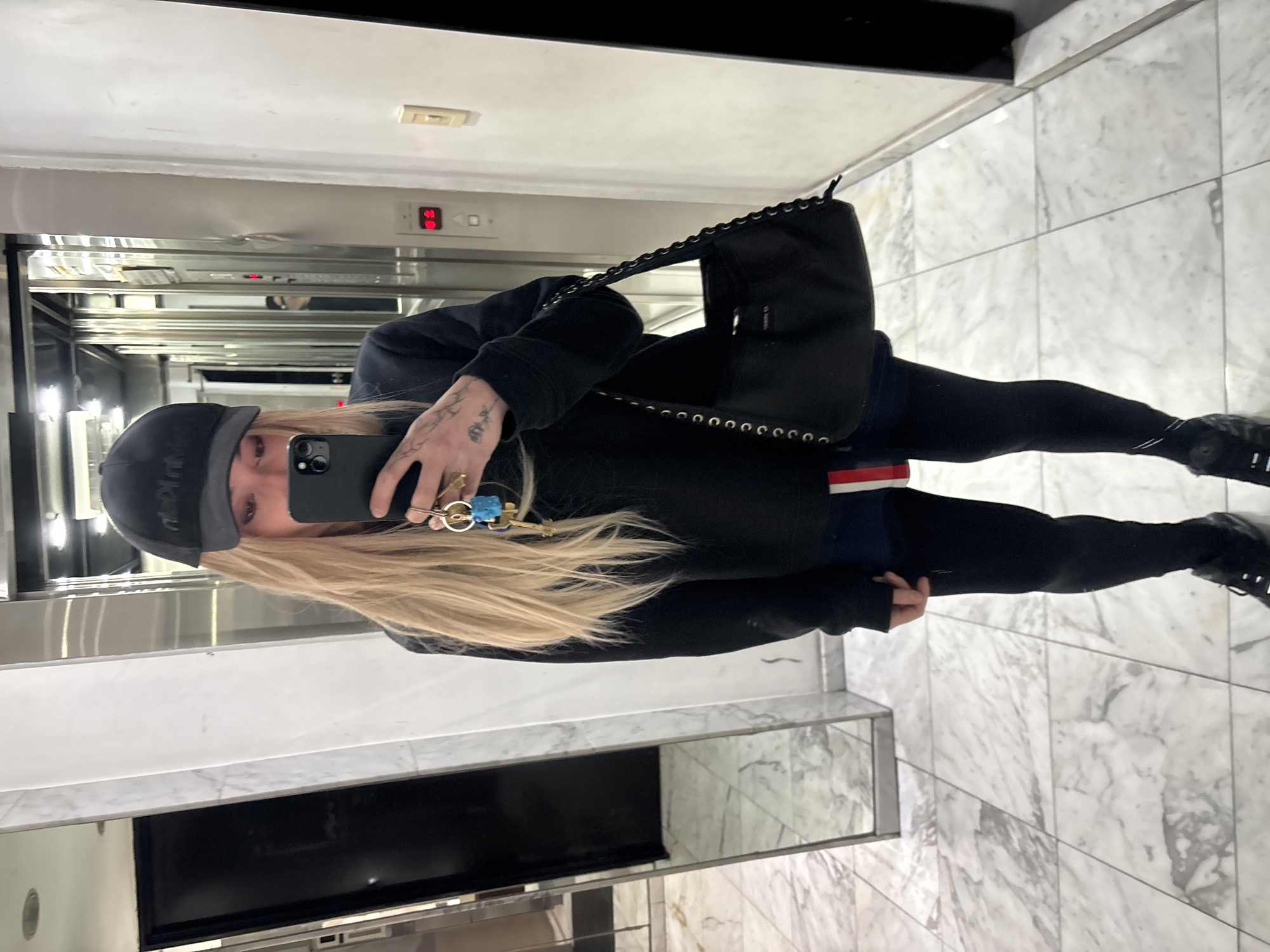 A person with long blonde hair, wearing a black baseball cap, black jacket, black leggings, and black shoes, stands inside an elevator, taking a selfie in the mirror. The person is holding a black smartphone in their left hand, with a set of keys hanging from it. A black shoulder bag with a chain strap is slung across their body. The elevator interior is primarily stainless steel, with a digital display showing the number "06" above the door. The floor outside the elevator is covered with white and grey marble tiles. The location appears to be inside a building in Buenos Aires, Argentina, but there are no specific identifying elements to confirm this.