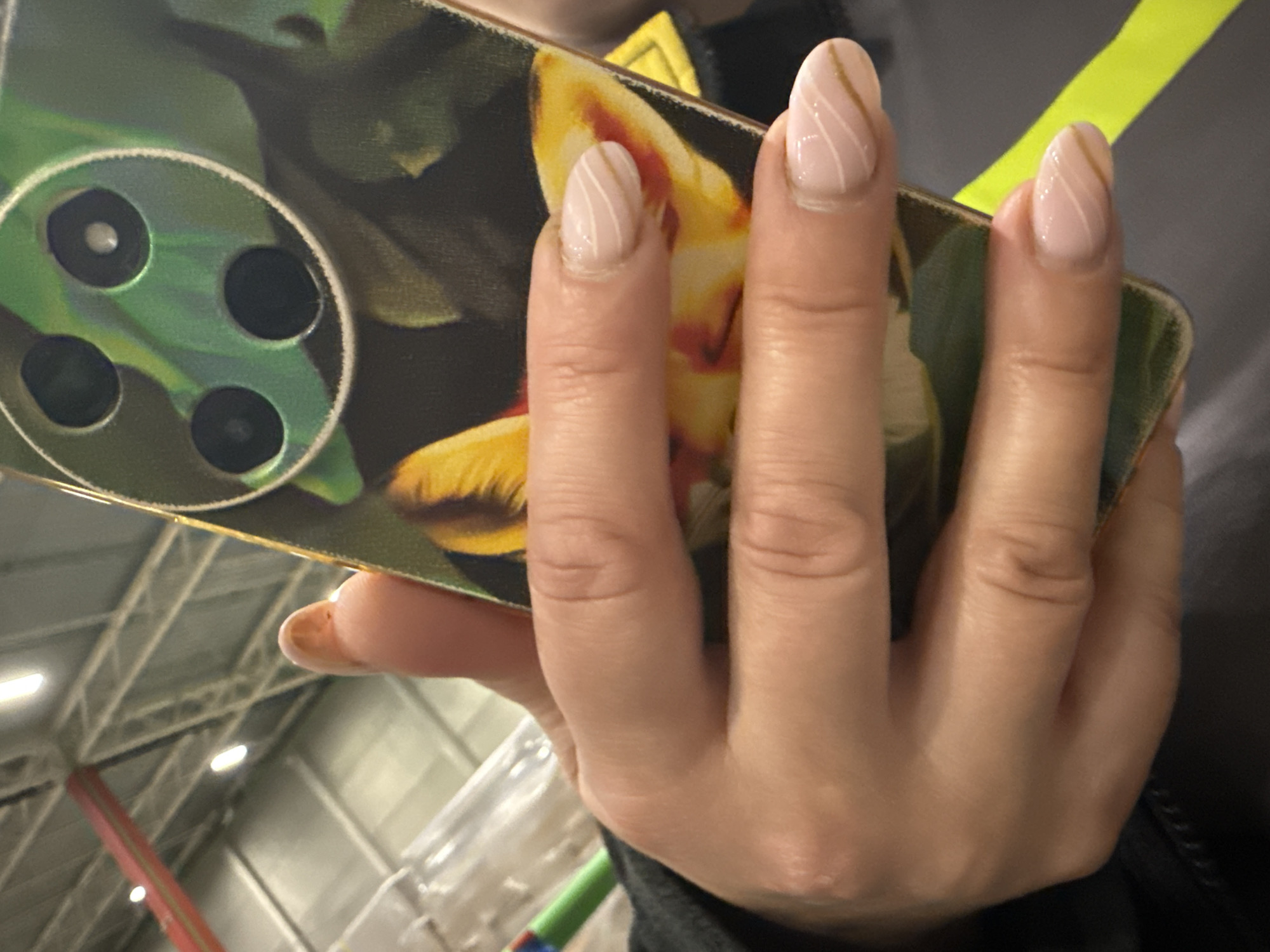 A hand holds a smartphone with a floral design phone case. The hand is slightly angled towards the viewer, displaying manicured nails with a nude base and a swirling white line design. The phone case features a printed image of a flower, possibly a tulip, in shades of yellow, orange, and green. The phone's camera lens array is visible, showing four lenses arranged in a circular configuration. The background shows parts of a building, including ceiling structures and interior elements like a colorful structure. The location is not specified or discernible from the image.
